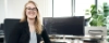 Eine Frau sitzt an einem Schreibtisch mit einem Tablet vor einem Computerbildschirm im Büro.