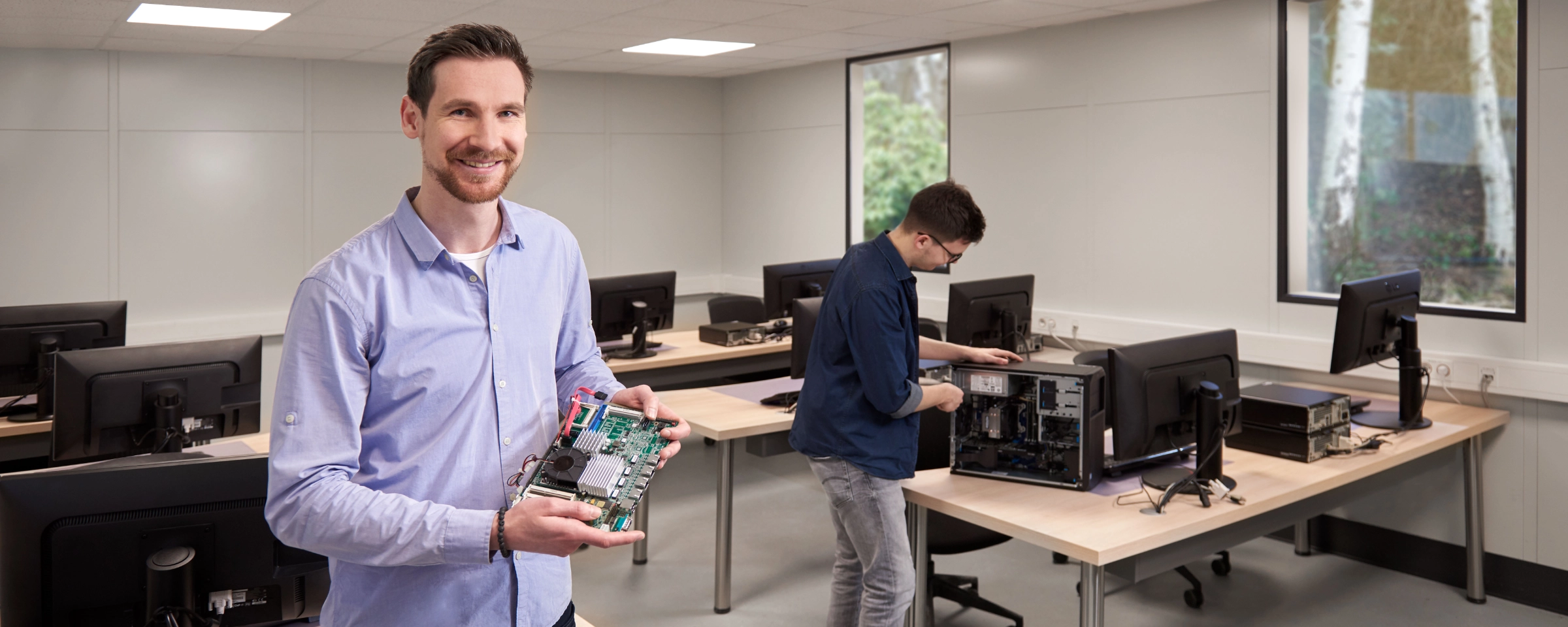 Ein Mann hält ein Computerteil in einem Klassenzimmer mit mehreren Computern und einem anderen Mann im Hintergrund.