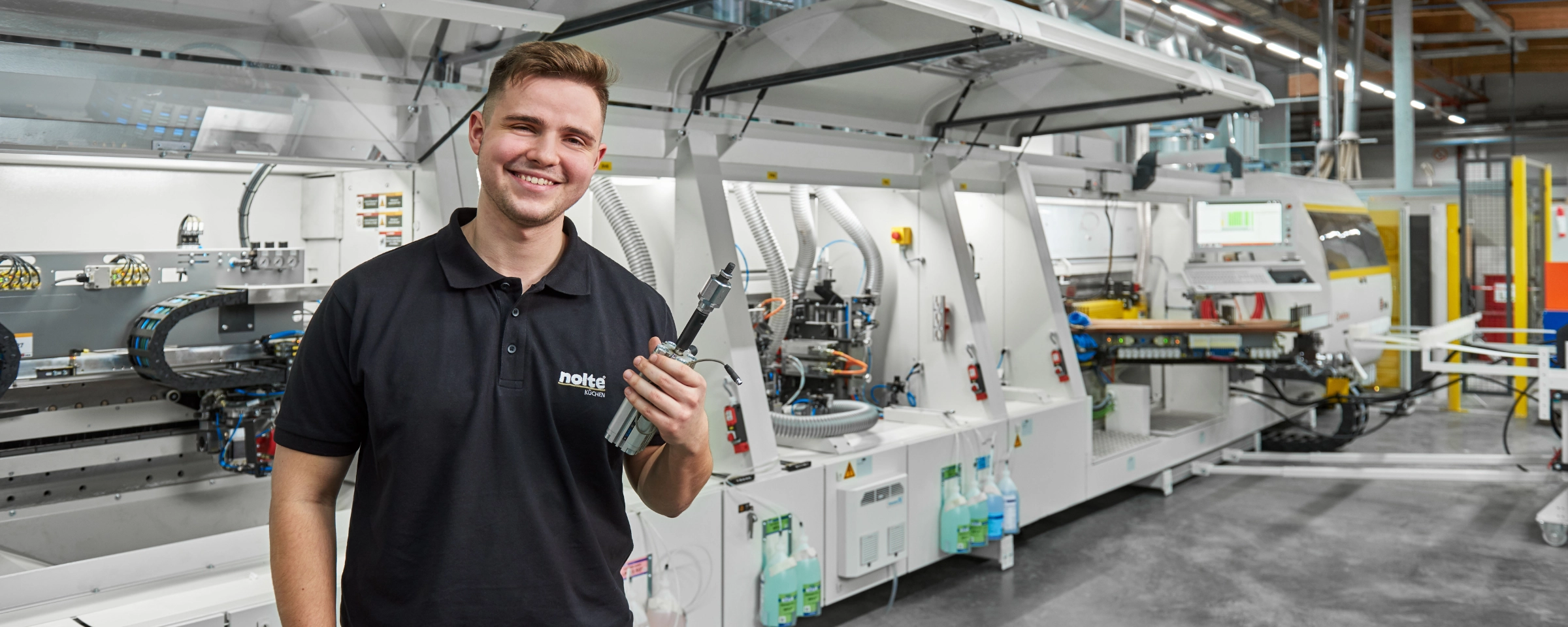 Ein lächelnder Mann steht in einer modernen Produktionshalle neben einer Maschine.
