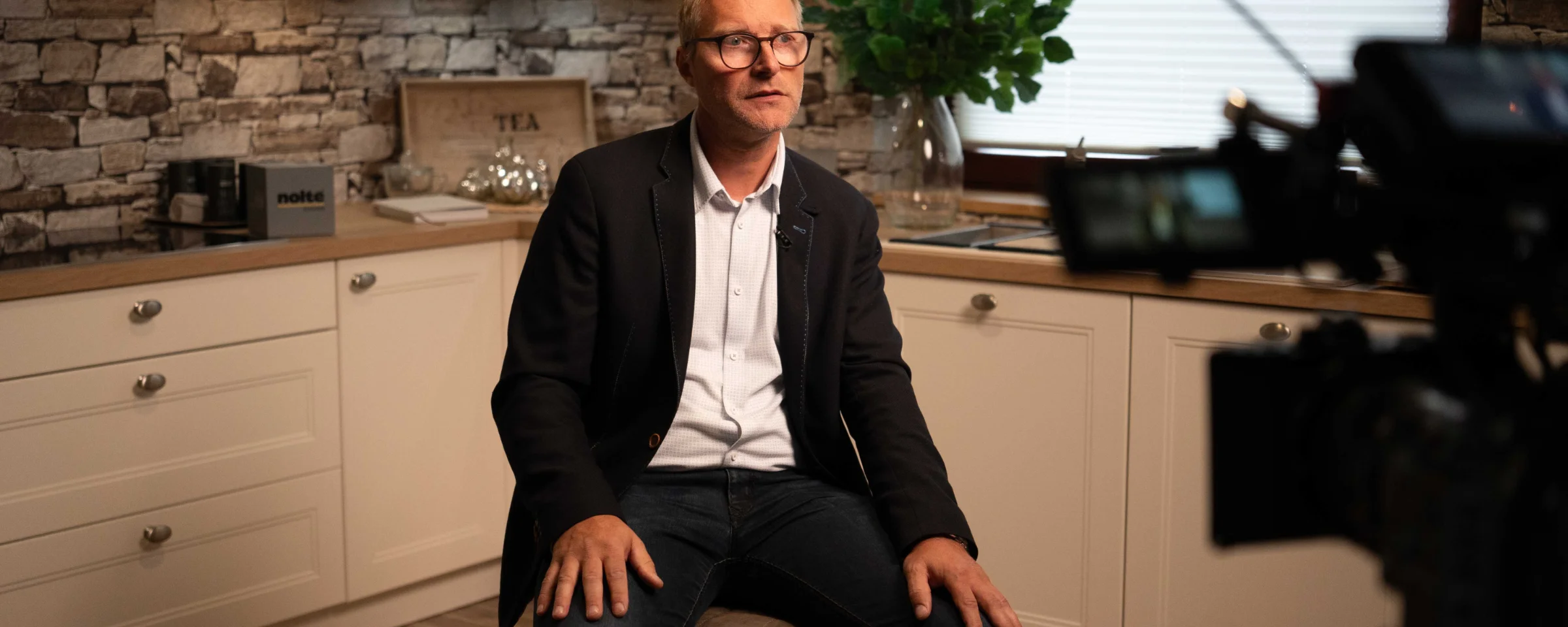 Un homme est assis dans une cuisine moderne avec mur en pierre et armoires blanches. Nolte Kitchen.