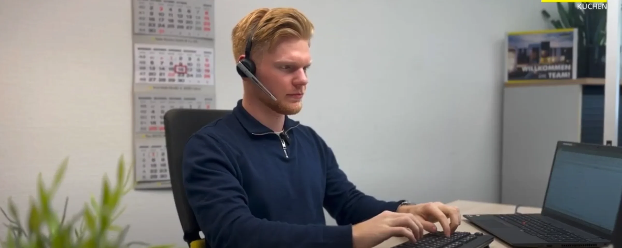 Ein Mann arbeitet konzentriert an einem Schreibtisch mit Computer und Headset; im Hintergrund das Logo von Nolte Küchen.