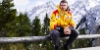 Un uomo con una giacca gialla seduto su una staccionata di legno davanti a montagne innevate.