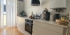 La cuisine moderne Nolte avec le programme SIGMA LACK, se trouve dans une pièce lumineuse avec de grandes fenêtres et des rideaux blancs du sol au plafond. Les armoires et tiroirs blancs sans poignées s'harmonisent avec le plancher en bois chaud. Un four encastrable noir avec plaque de cuisson est placé au centre, à côté un évier avec un robinet noir. Sur le plan de travail, qui a été intégré des deux côtés par le client, se trouvent une machine à espresso, une carafe d'eau noire et un bac à compost. Une lampe suspendue noire ajoute une touche moderne. Des détails personnels comme un panneau «ON STAGE» et un plateau Skyline de Frankfurt complètent l'ambiance.