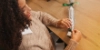 Eine Frau sitzt an einem Holztisch und bindet ein Geschenkband um ein verpacktes Geschenk.
