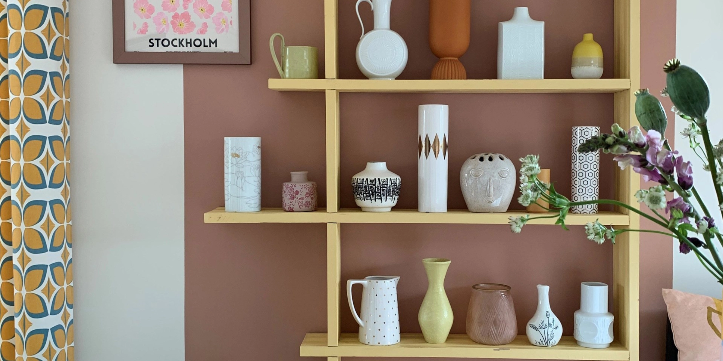 A shelf with vases and decor against a pink wall, with a cushion and magazine in the foreground. Nolte Living.