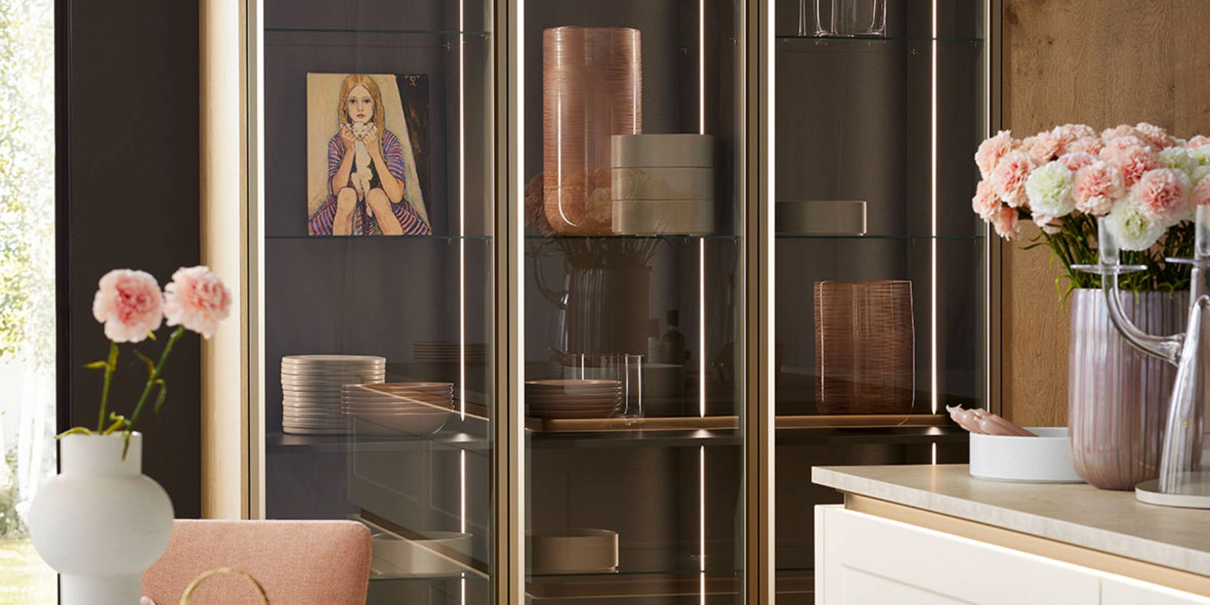 Une cuisine moderne avec vitrines en verre et façades en bois, décorée de fleurs et d'art, par Nolte Kitchen.