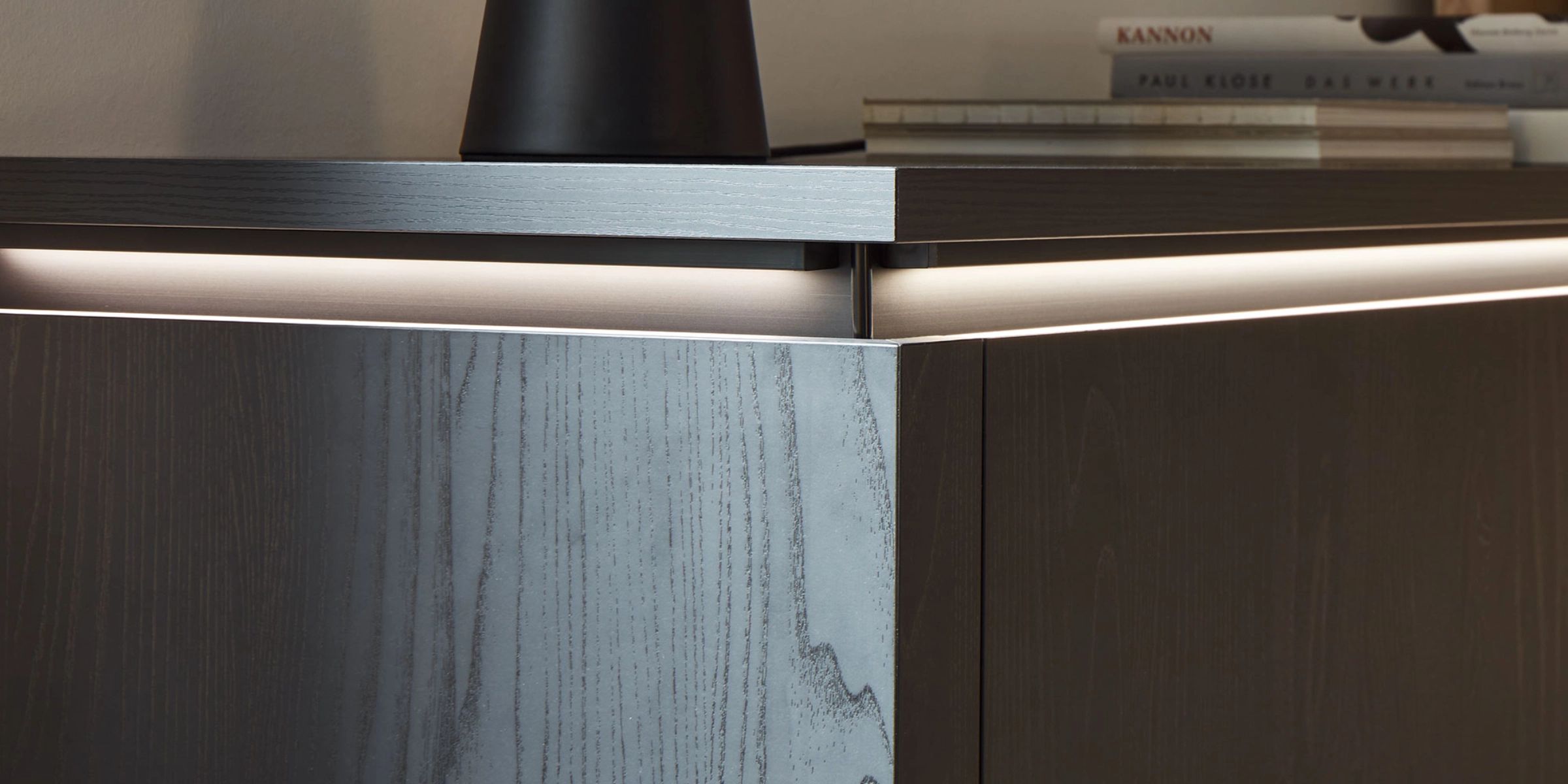 Un buffet moderne en bois foncé avec éclairage, livres et décorations dessus, par Nolte Living.