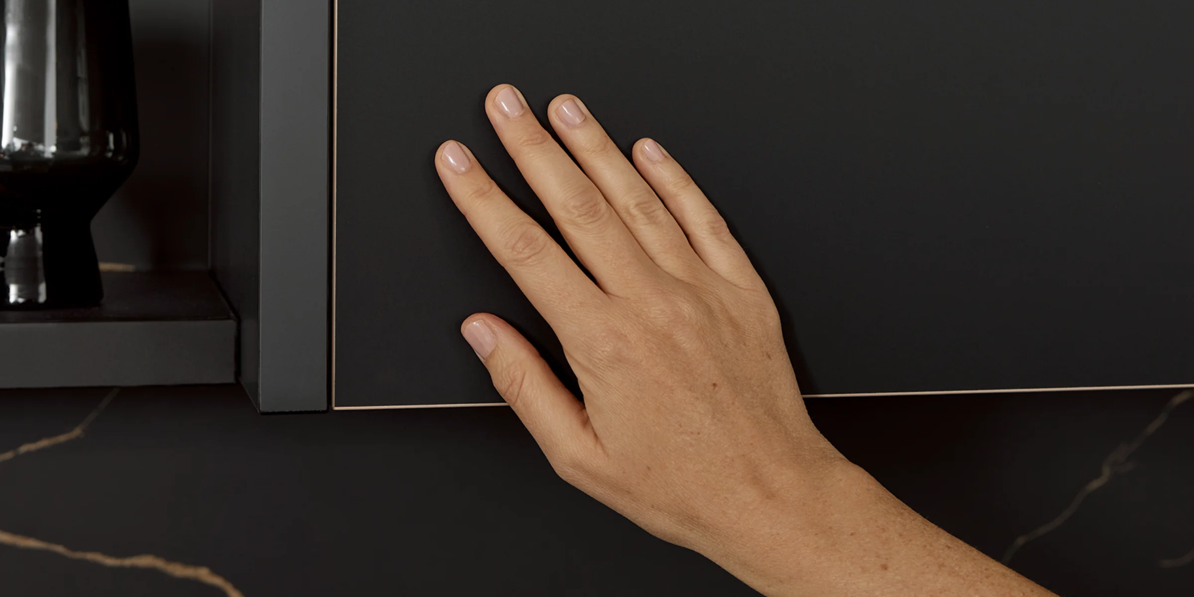 La main touche une façade de cuisine noire mate FLAIR avec un bord raffiné aspect laiton, dotée d'une surface anti-traces de doigts.
