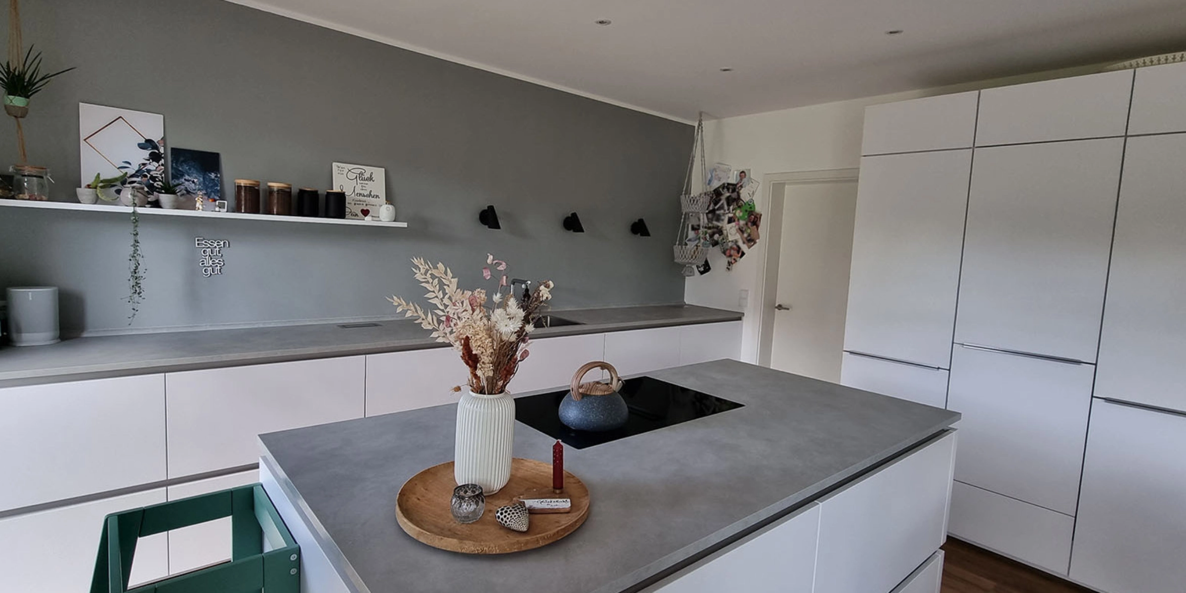 Moderne, minimalistische Nolte keuken van een Nolte Küchen medewerkster met een betonnen werkblad en fronten uit het programma PLUS in Arcticweiß. In het midden bevindt zich een keukeneiland met een inductiekookplaat en een vaas met droogbloemen. Aan de wand hangt een open rek met decoratie, glazen en een mintgroen koffiezetapparaat. Een magneetbord met foto's en hangmandjes geeft de keuken een persoonlijk accent. De keuken oogt opgeruimd, stijlvol en functioneel.