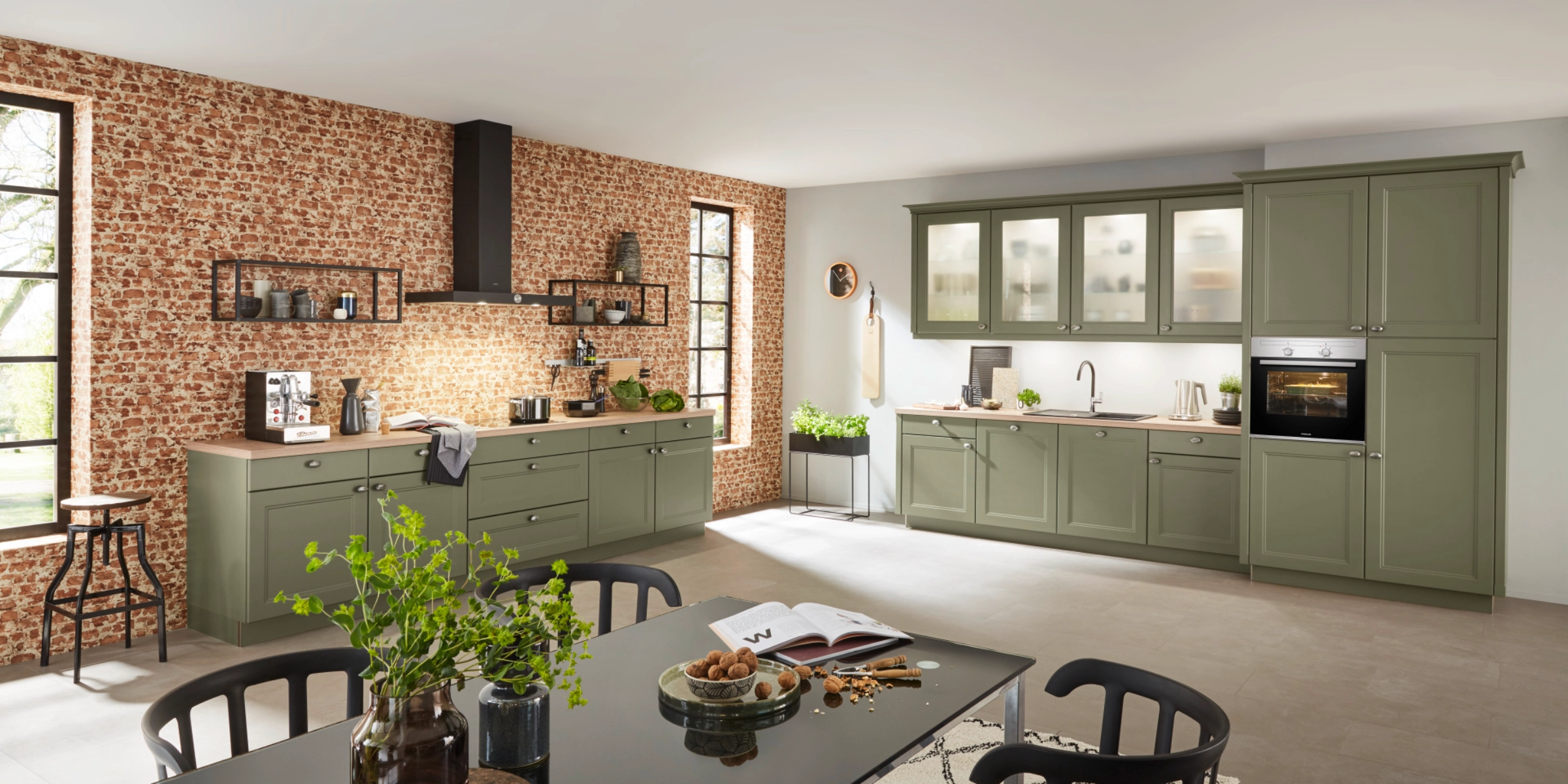Cucina in stile country di Nolte con la frontale WINDSOR LACK in oliva softmatt. La colorazione naturale è completata da un piano di lavoro in quercia di pietra. Scaffali a muro neri in stile industriale creano contrasti moderni con l'aspetto classico della cornice delle frontali. La cucina unisce il comfort rurale con la chiarezza urbana – funzionale, accogliente e stilosa.