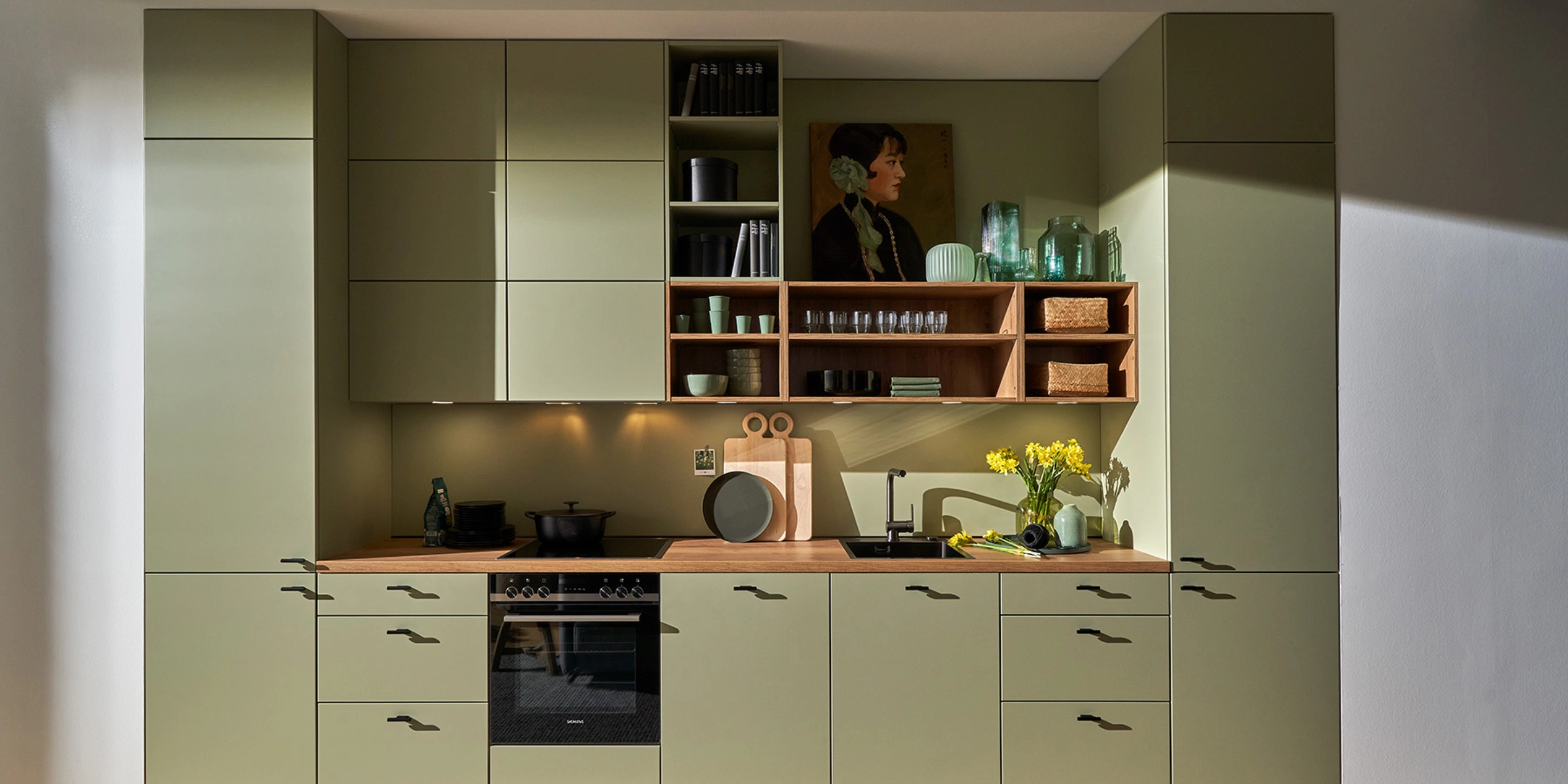 Cuisine Nolte élégante avec la façade SOFT LACK en Avocado softmatt. La ligne de cuisine sans poignées combine un vert doux avec un plan de travail en bois chaud et des étagères ouvertes. L’atmosphère conviviale est soulignée par des éléments décoratifs, des plantes et des matériaux naturels – une cuisine moderne avec des couleurs fraîches et des lignes épurées.