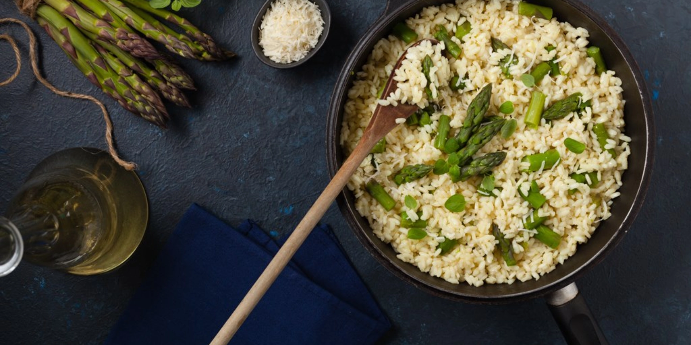 Padella con risotto agli asparagi, asparagi freschi e un bicchiere di vino bianco su sfondo scuro.