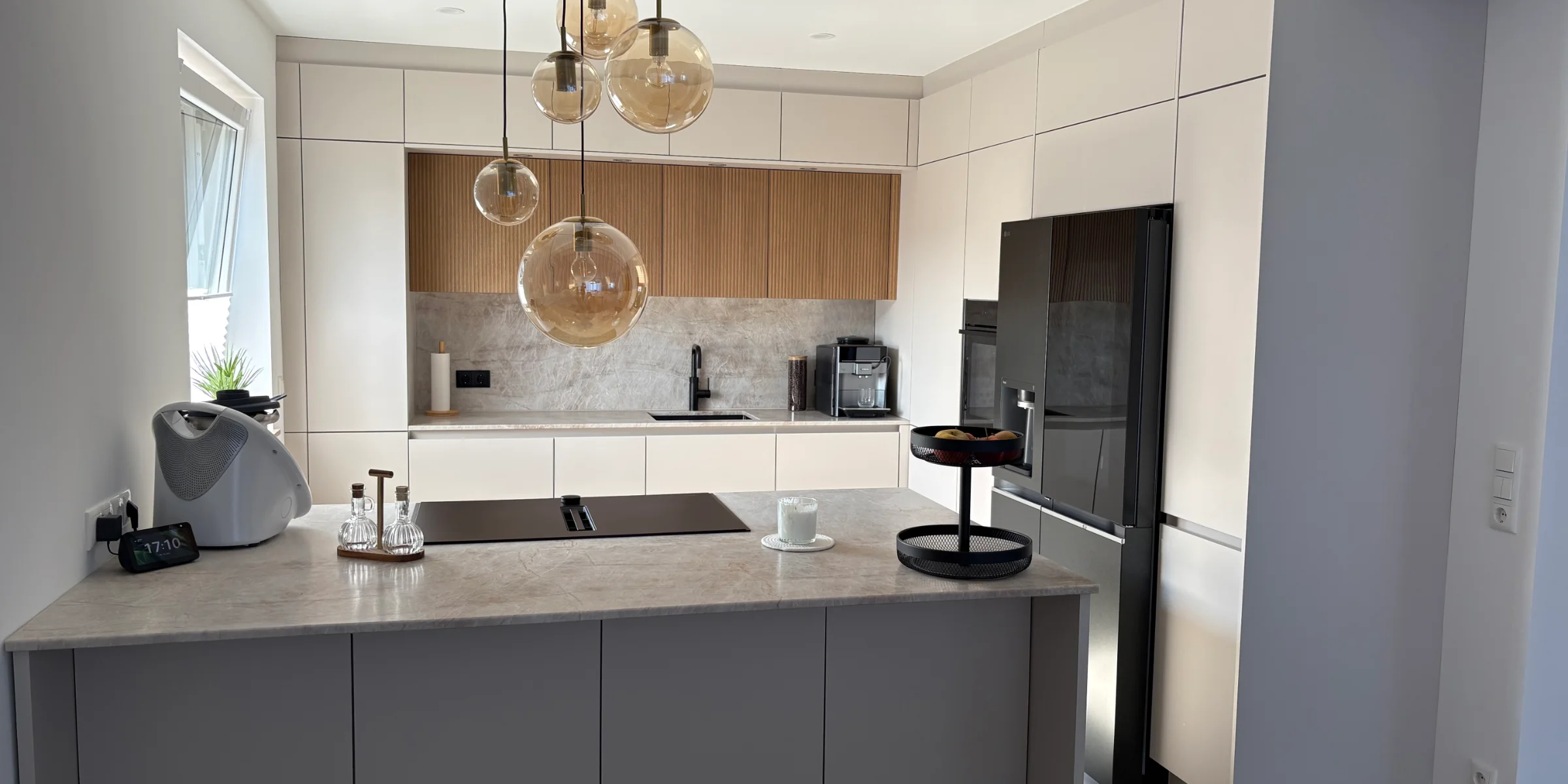 Nowoczesna kuchnia marki Nolte z wyspą kuchenną, frontami w wykończeniu SOFT LACK Sahara w matowym odcieniu oraz neoLODGE dąb Diamant, szafkami sięgającymi sufitu, czarną armaturą oraz jasnym wykończeniem imitującym naturalny kamień w otwartej aranżacji przestrzeni dziennej.