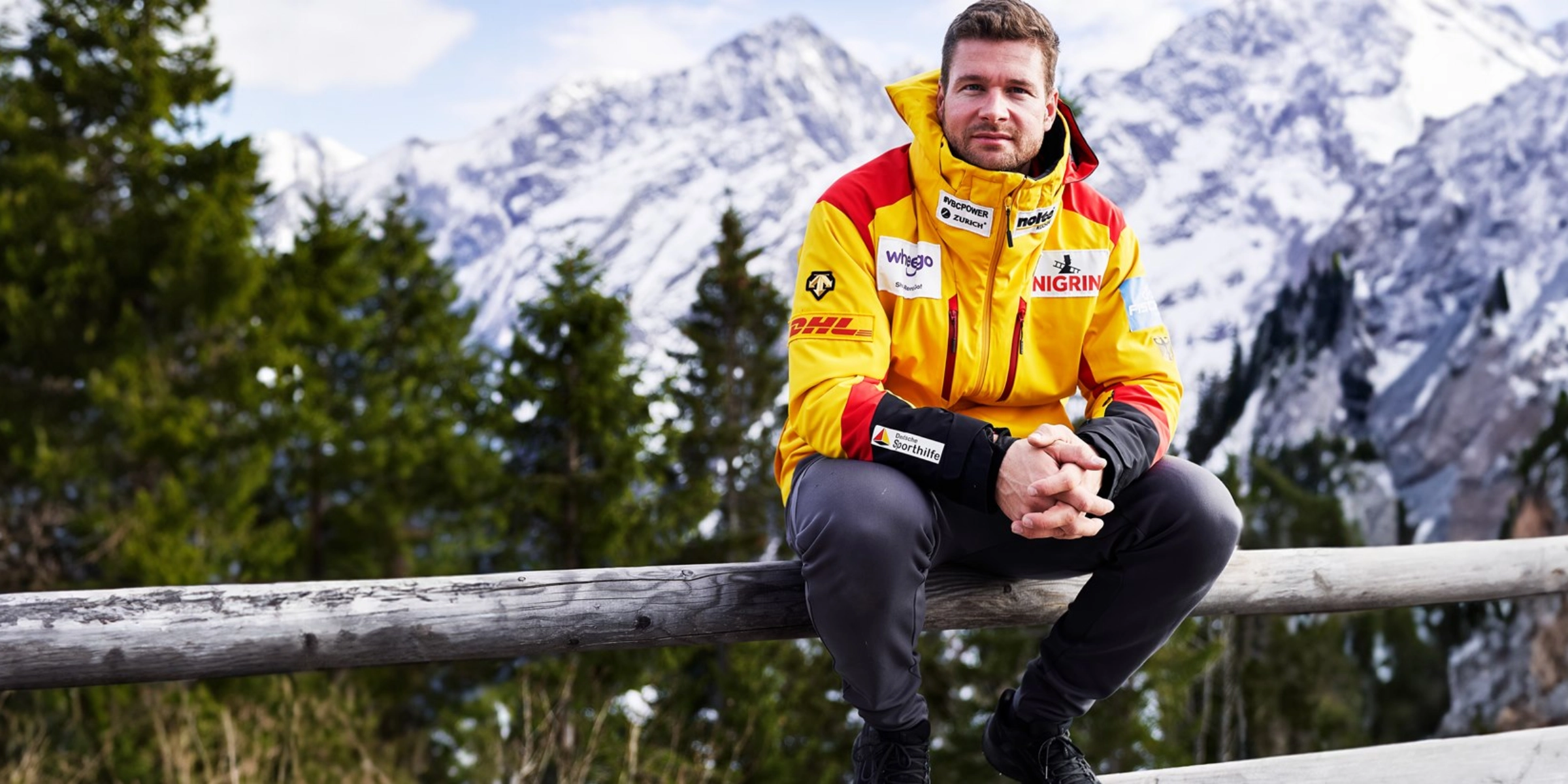 Un uomo con una giacca gialla seduto su una staccionata di legno davanti a montagne innevate.
