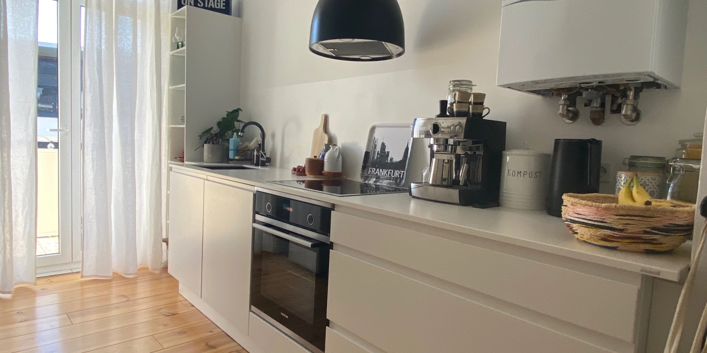 La cuisine moderne Nolte avec le programme SIGMA LACK, se trouve dans une pièce lumineuse avec de grandes fenêtres et des rideaux blancs du sol au plafond. Les armoires et tiroirs blancs sans poignées s'harmonisent avec le plancher en bois chaud. Un four encastrable noir avec plaque de cuisson est placé au centre, à côté un évier avec un robinet noir. Sur le plan de travail, qui a été intégré des deux côtés par le client, se trouvent une machine à espresso, une carafe d'eau noire et un bac à compost. Une lampe suspendue noire ajoute une touche moderne. Des détails personnels comme un panneau «ON STAGE» et un plateau Skyline de Frankfurt complètent l'ambiance.