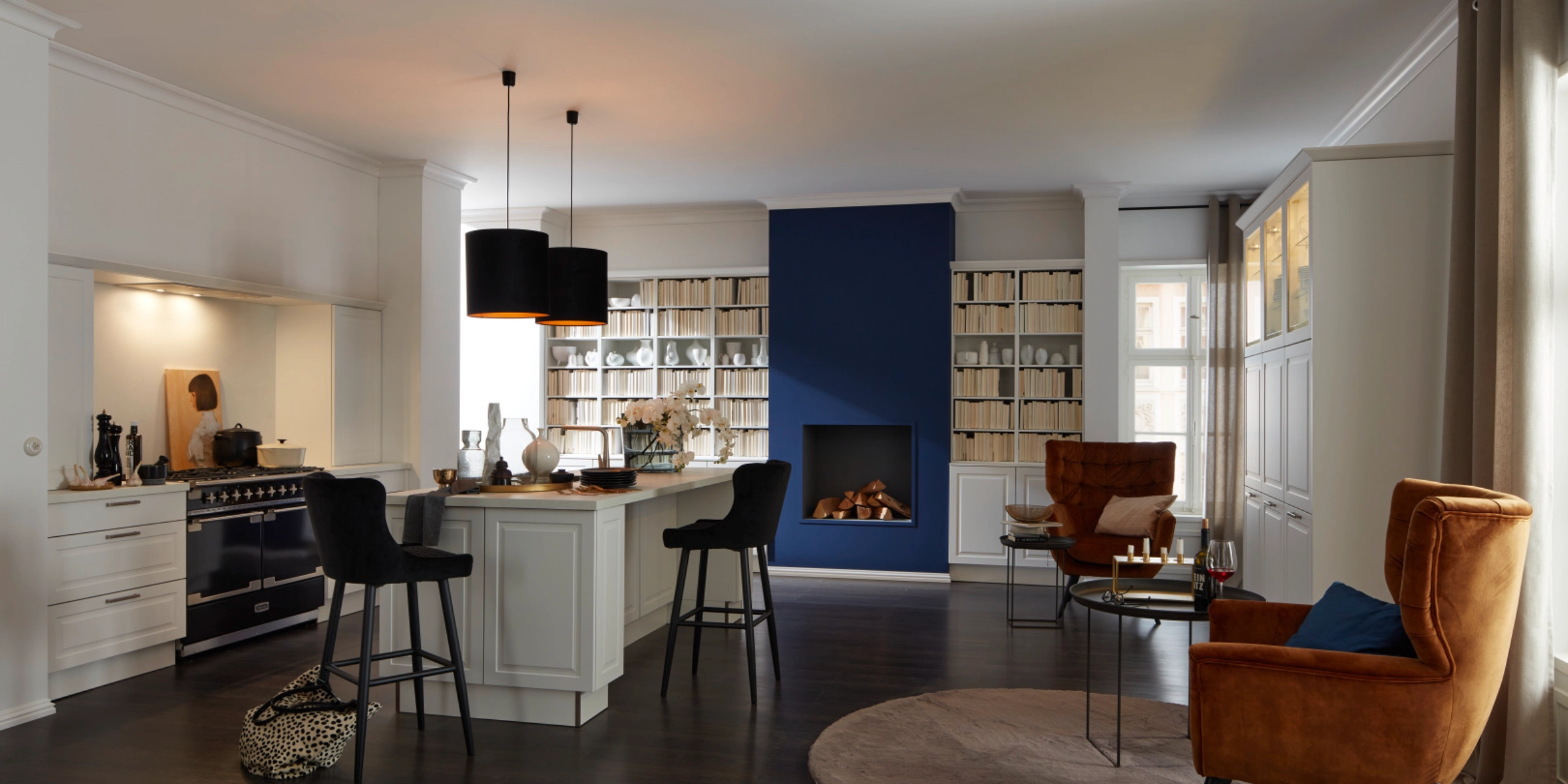 Cuisine Nolte conviviale de style maison de campagne avec la façade PORTO blanc softmatt. La cuisine séduit par son aspect à cadre classique, une zone de cuisson noire de style maison de campagne et un espace îlot de cuisson avec des tabourets de bar. Les façades claires s’harmonisent avec le sol en bois foncé, tandis qu’un espace de vie intégré avec cheminée, étagères à livres et fauteuils lounge en velours couleur cognac crée une ambiance élégante et chaleureuse.