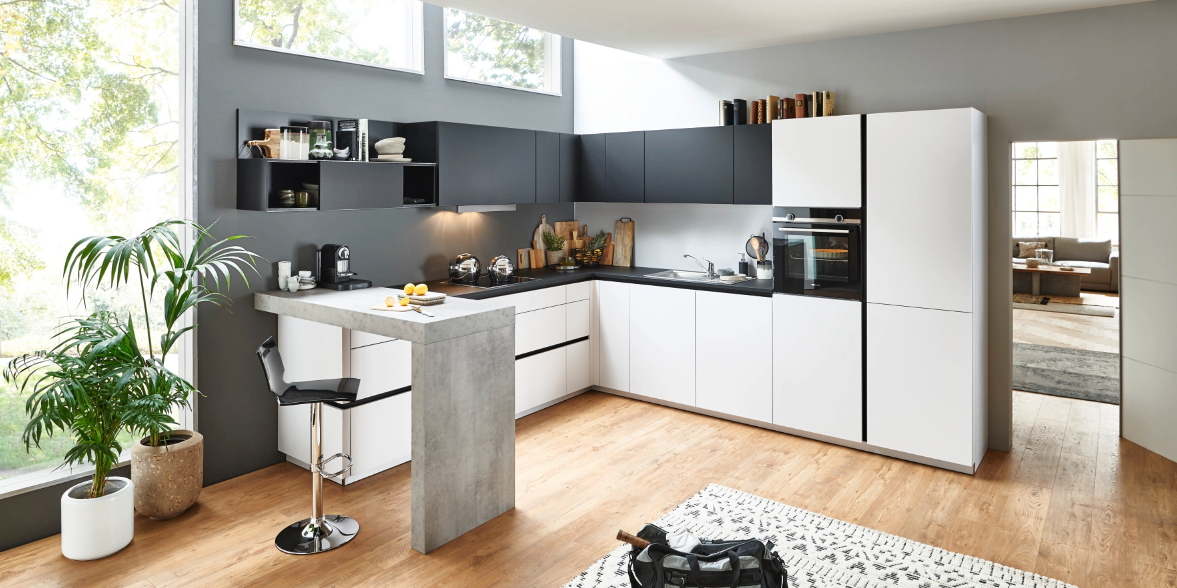 Cuisine Nolte moderne en forme de L et bar avec façades FEEL en Arcticweiß softmatt et façades PLUS en Schwarz softmatt. Le design sans poignées est complété par un comptoir marquant en aspect béton. L'effet lumineux et ouvert de la pièce est soutenu par beaucoup de lumière naturelle et des matériaux naturels comme le parquet en bois et les plantes – un espace élégant et fonctionnel pour cuisiner et vivre.