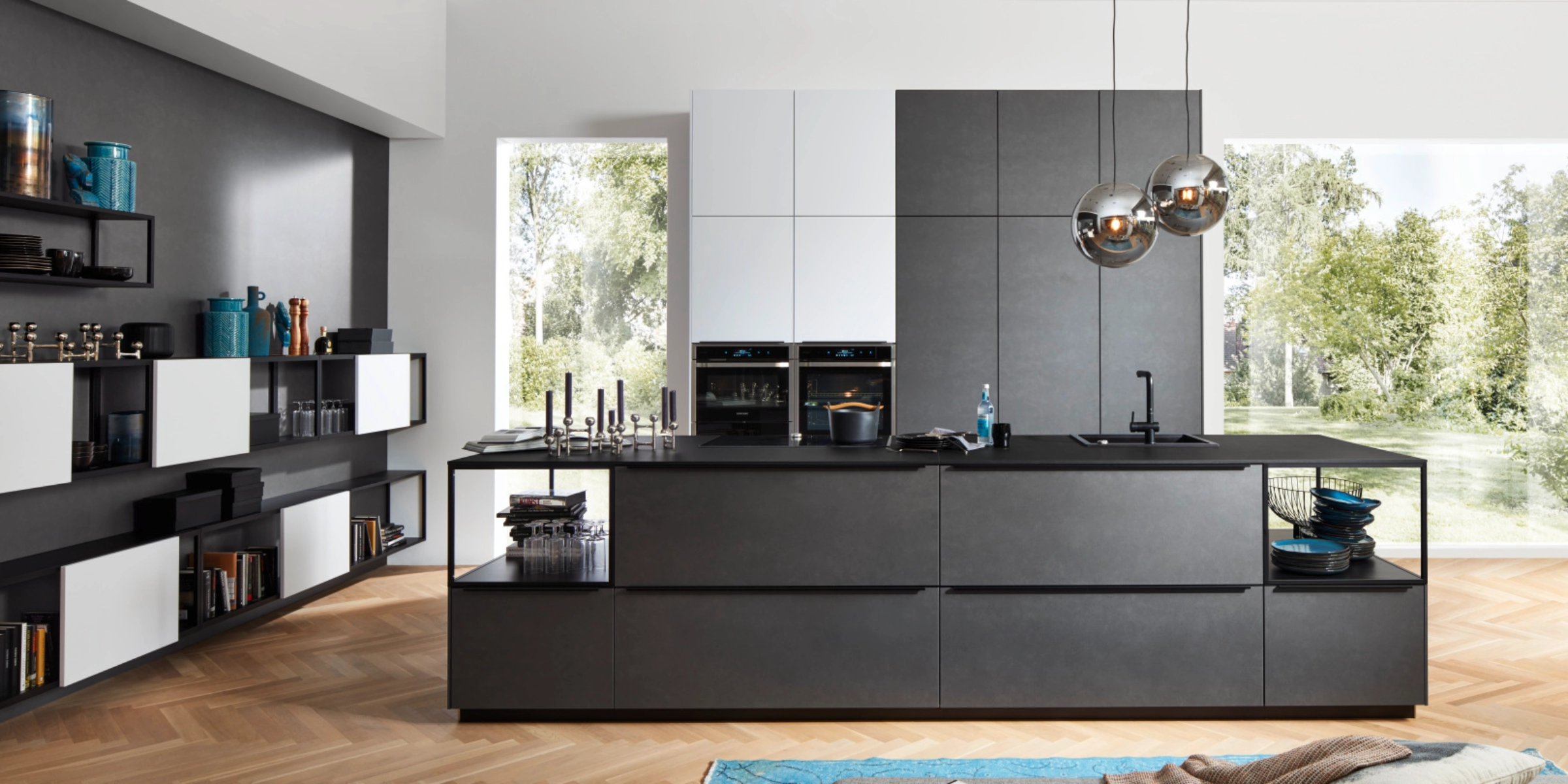 Cuisine Nolte moderne avec la façade FERRO en acier bleu et des armoires murales contrastantes en SOFT LACK blanc arctique soft mat. L’îlot sans poignées séduit par des lignes claires, des étagères ouvertes et des solutions de rangement intégrées. De grandes fenêtres offrent une ambiance baignée de lumière, qui souligne avec style le caractère industriel des surfaces en acier bleu.