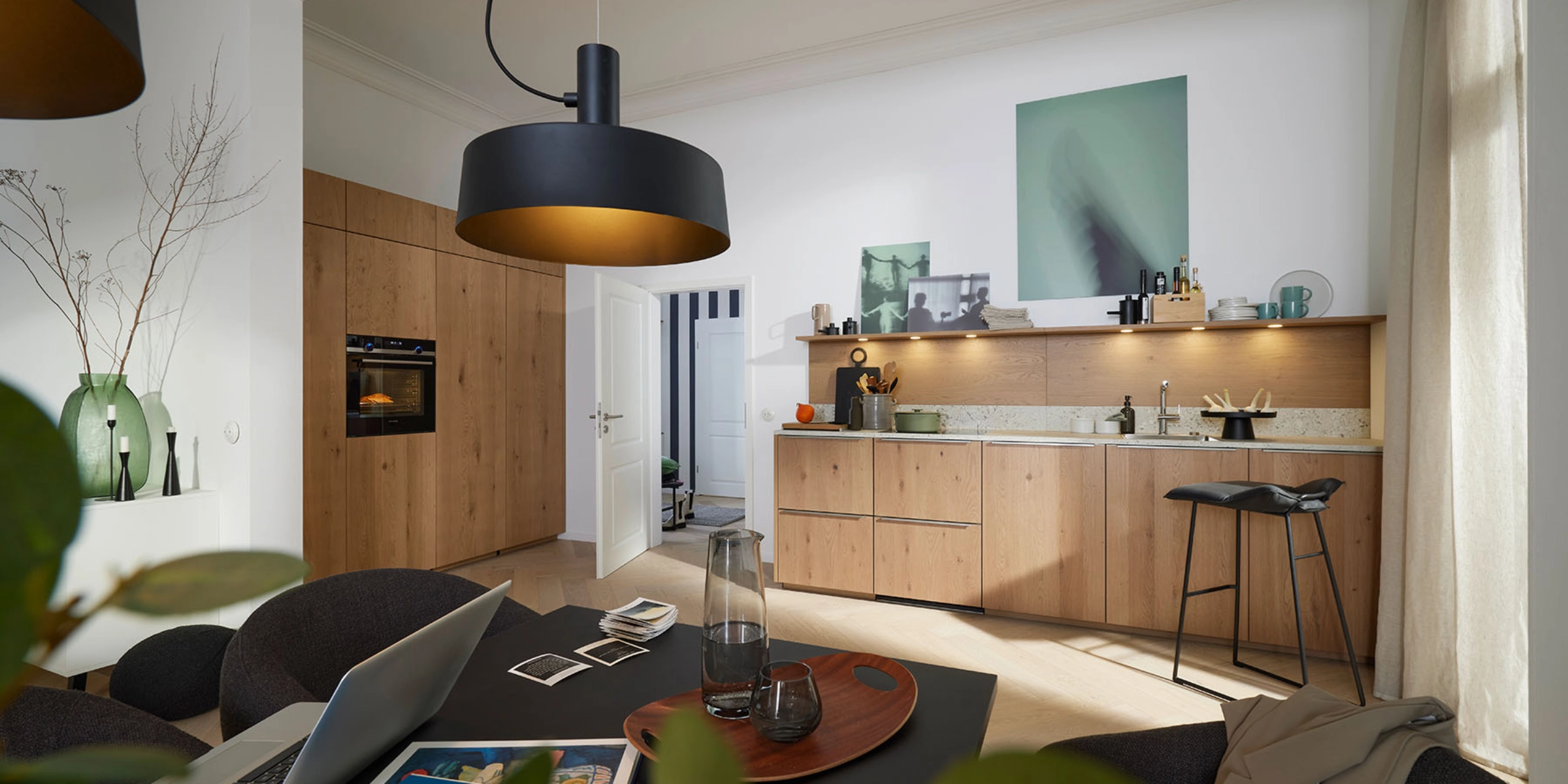 Cuisine moderne avec façades en bois et éclairage élégant, table à manger au premier plan. Nolte Kitchen.