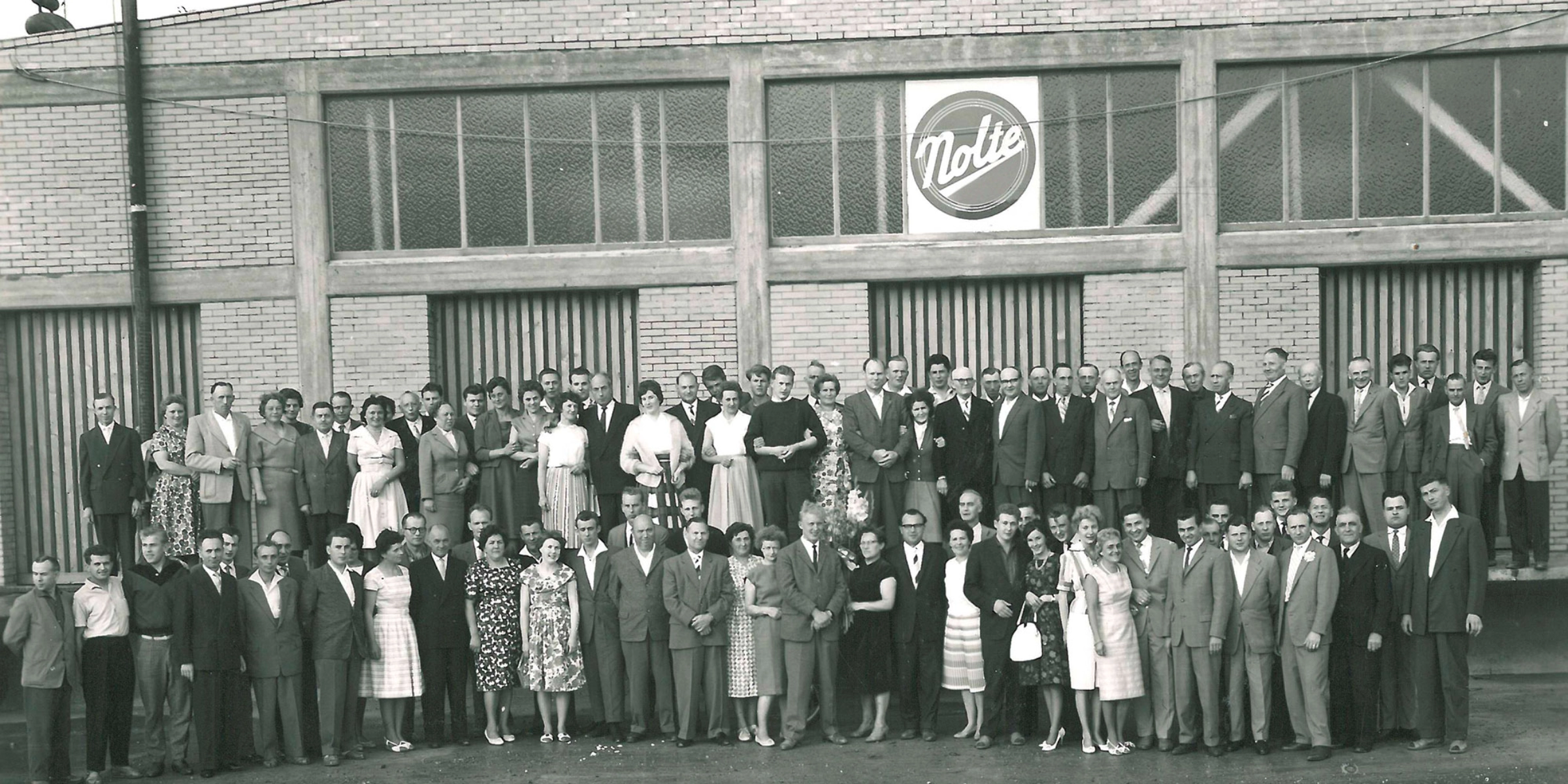 Un grand groupe de personnes se tient devant un bâtiment avec un logo "Nolte".