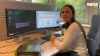 Eine Frau sitzt an einem Schreibtisch mit zwei Monitoren in einem Büro, links ein Buch über IT.