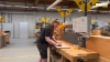 In einer Werkstatt arbeitet eine Person an einem Holztisch mit Werkzeugen; im Hintergrund sind weitere Arbeitsplätze zu sehen.