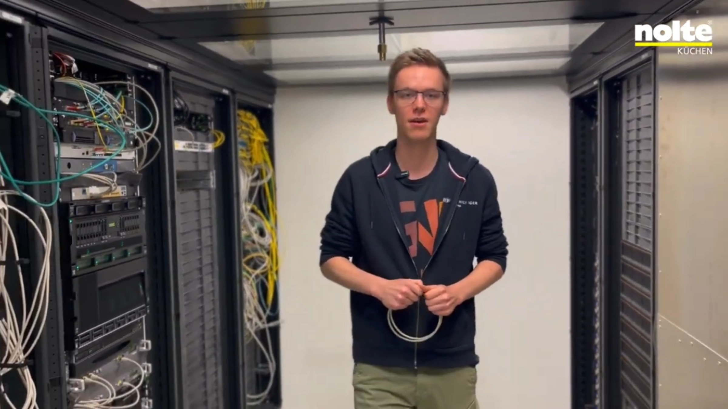 Ein Mann steht in einem Serverraum zwischen Regalen mit Kabeln und Technik.