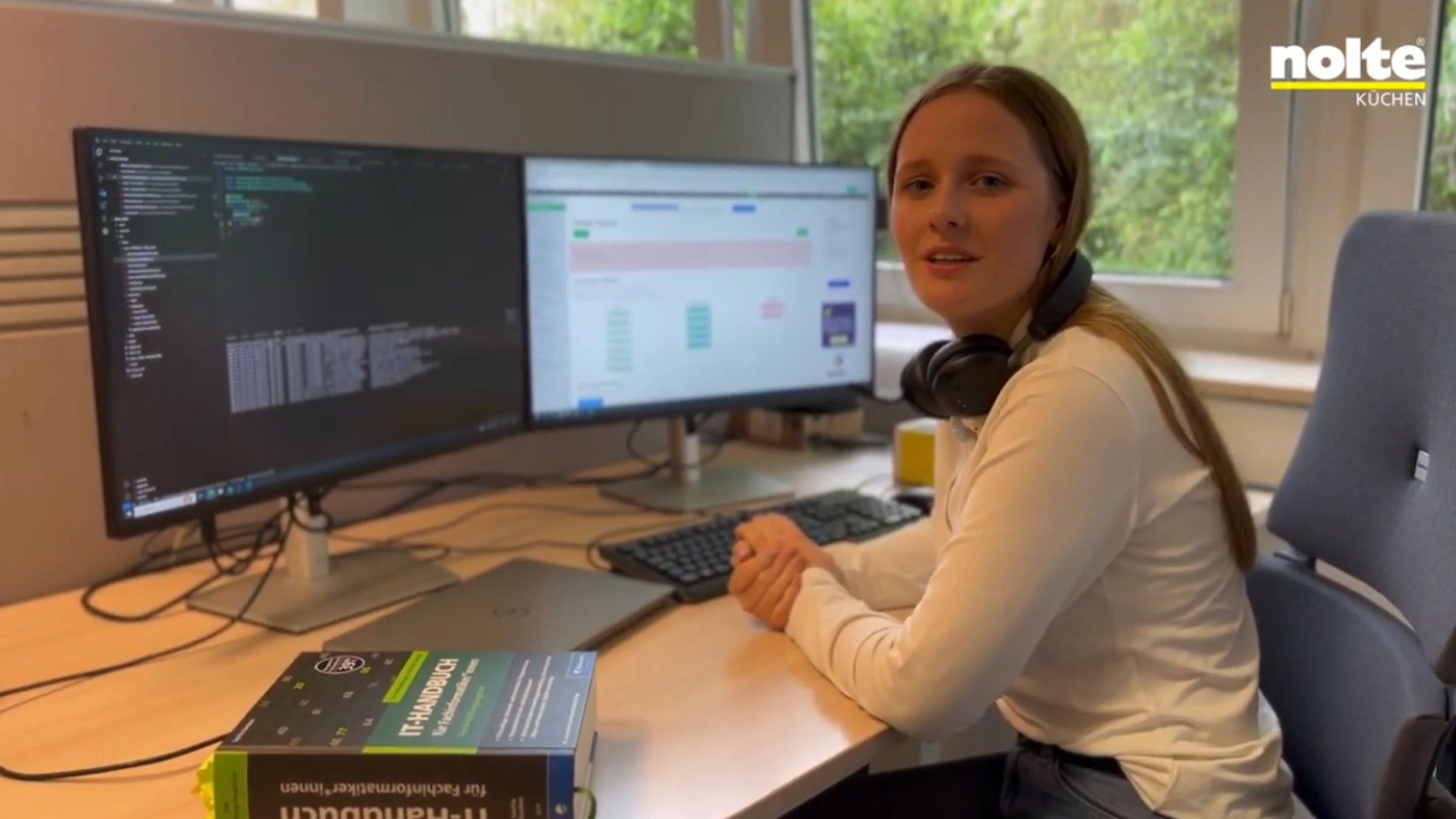 Eine Frau sitzt an einem Schreibtisch mit zwei Monitoren in einem Büro, links ein Buch über IT.