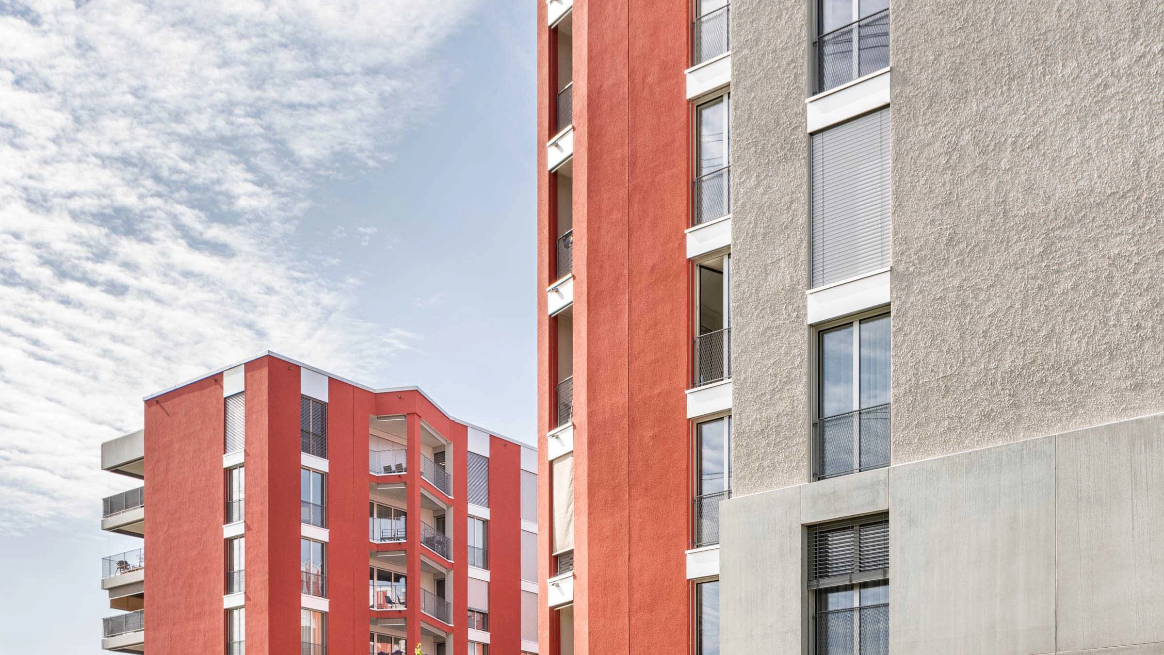 Zwei moderne Wohngebäude mit roter und grauer Fassade unter blauem Himmel.
