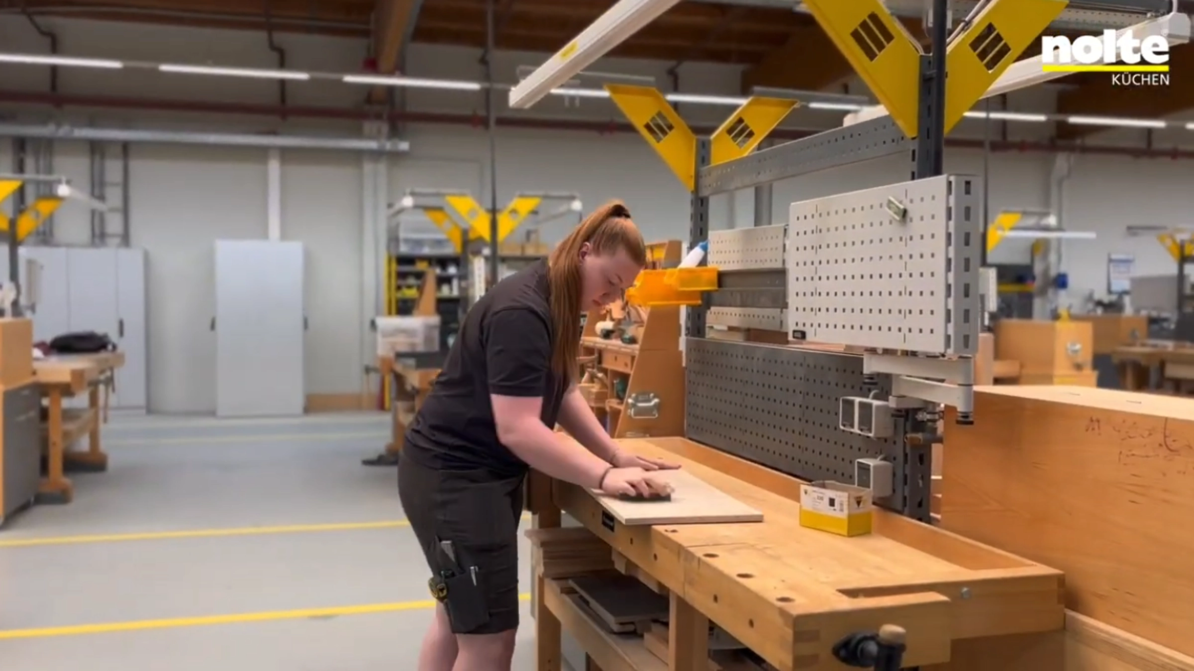 In einer Werkstatt arbeitet eine Person an einem Holztisch mit Werkzeugen; im Hintergrund sind weitere Arbeitsplätze zu sehen.