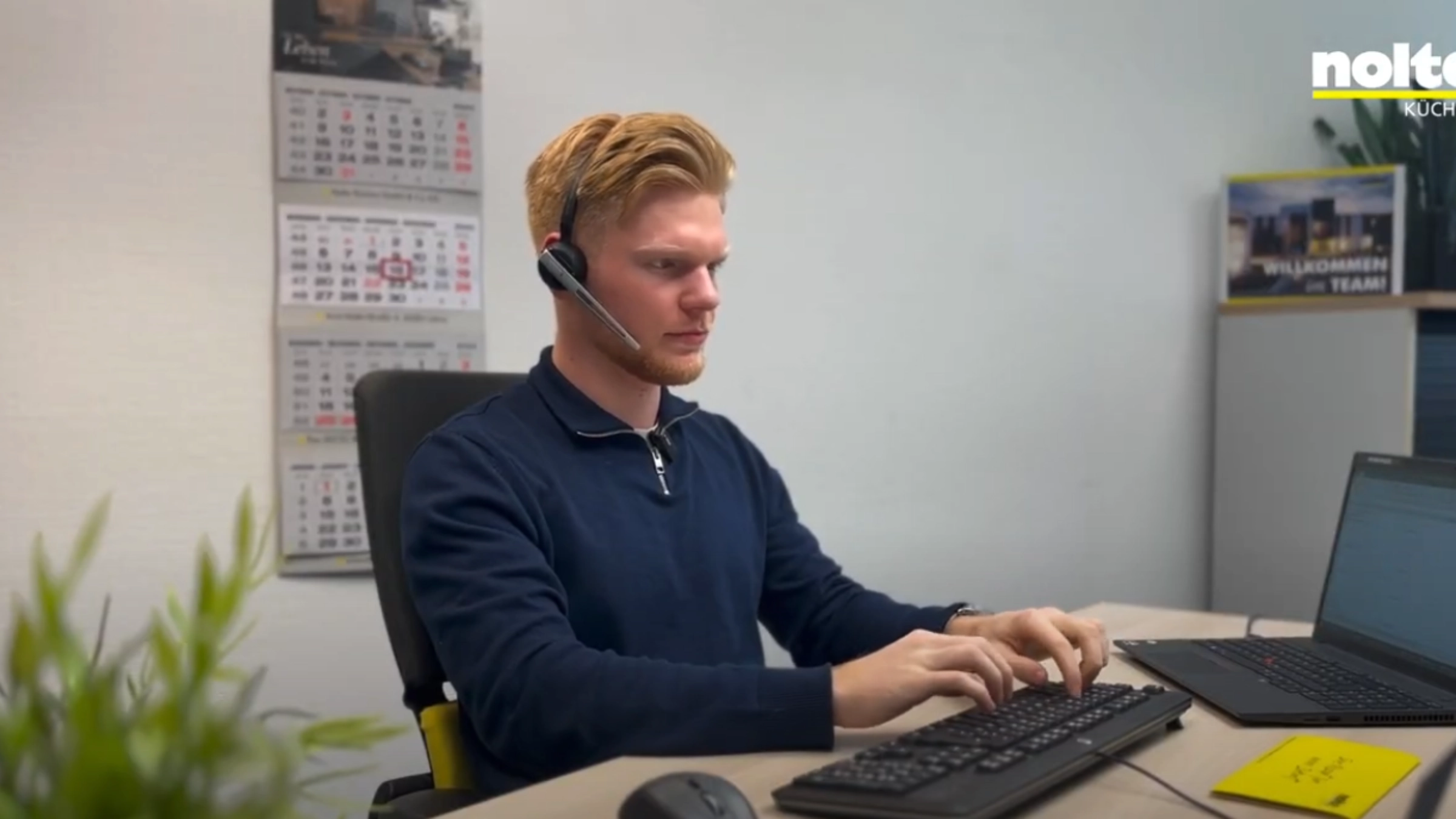 Ein Mann arbeitet konzentriert an einem Schreibtisch mit Computer und Headset; im Hintergrund das Logo von Nolte Küchen.