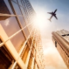 A plane flies over modern skyscrapers in a city at sunset.