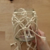 Une main tient un bocal décoré de macramé sur une table en bois.
