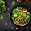 Assiette noire avec salade de quinoa, brocoli, champignons et feuilles de basilic sur table sombre.