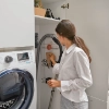 Une femme organise une buanderie avec aspirateur, machine à laver et étagères. Nolte buanderies.