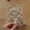 Une main tient un verre décoré de macramé sur une table en bois.