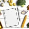 Un carnet avec un stylo entouré de légumes sur une table en bois blanc.
