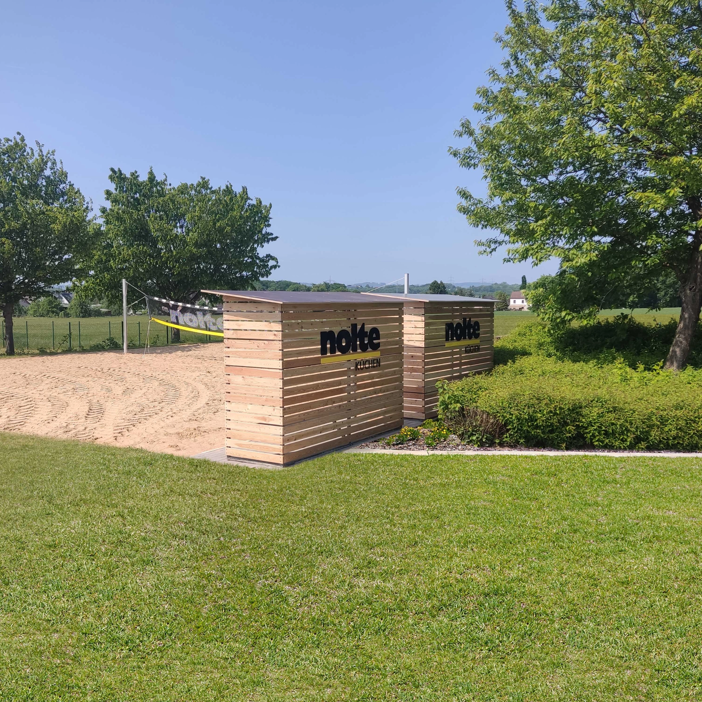 Een grasveld met bomen en een zandplaats, op de achtergrond borden met "Nolte Kitchen".