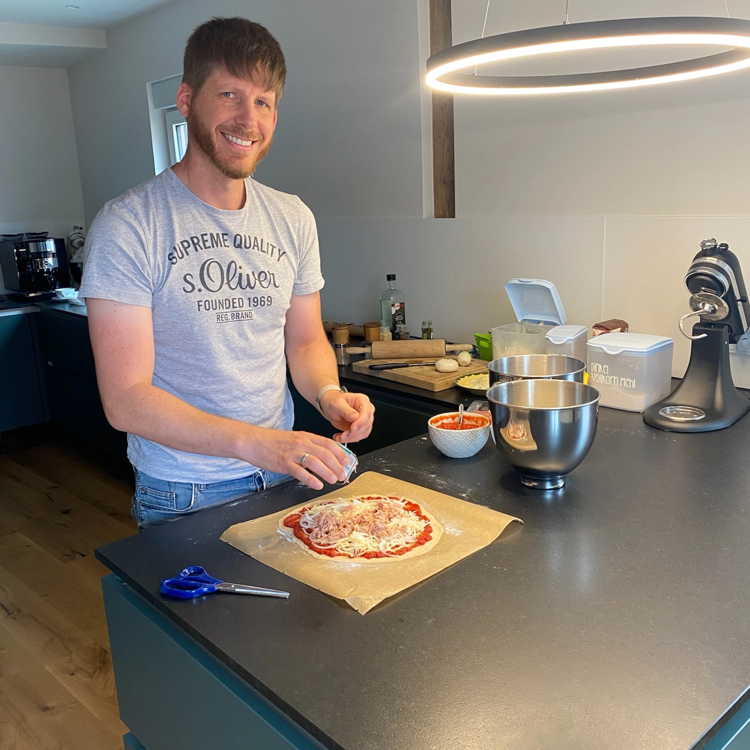 Un homme prépare une pizza dans une cuisine moderne, entouré d'appareils et d'ingrédients. Nolte Kitchen.
