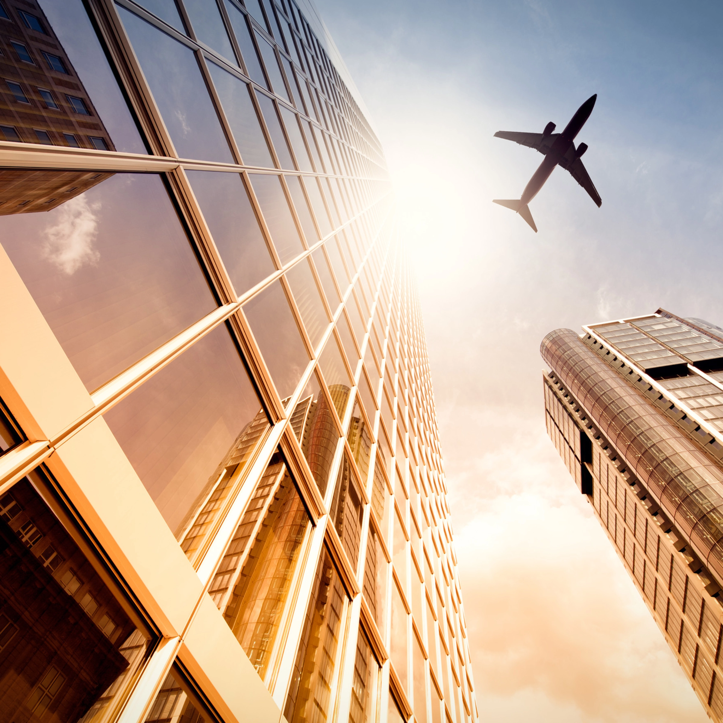A plane flies over modern skyscrapers in a city at sunset.