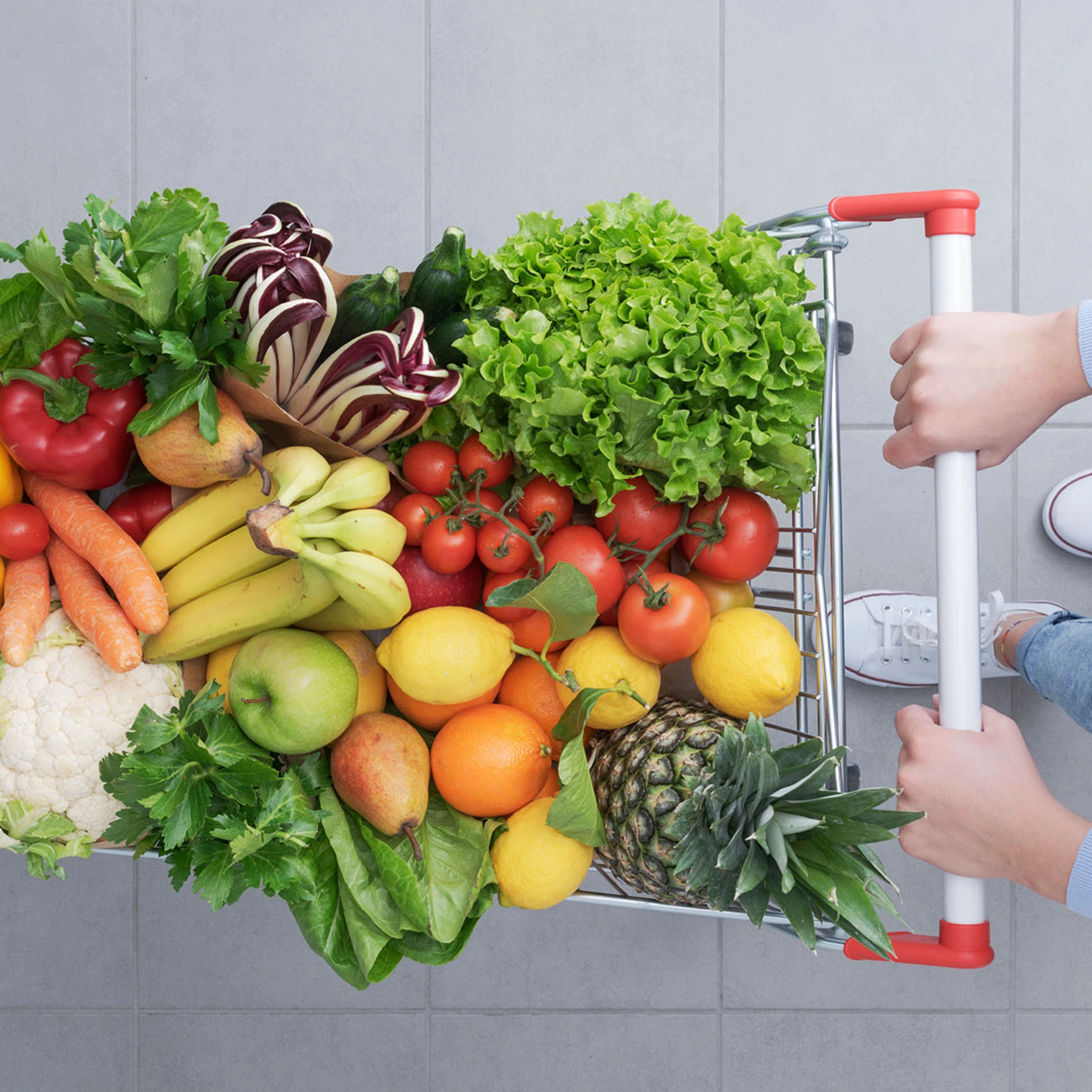 Iemand duwt een winkelwagentje vol vers fruit en groenten over een grijze tegelvloer.