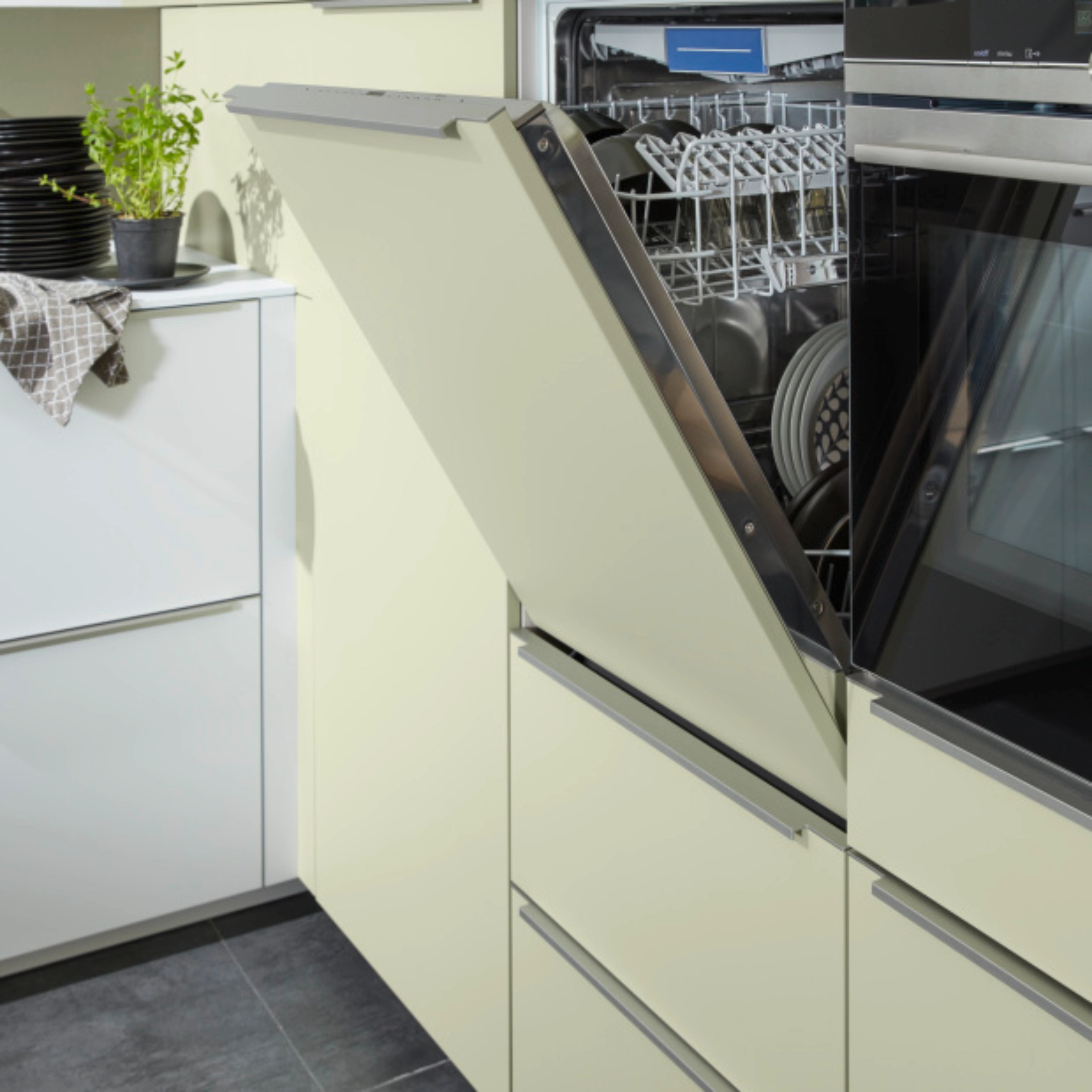 Un lave-vaisselle ouvert dans une cuisine moderne avec des armoires claires, des plantes et de la vaisselle. Nolte Kitchen.