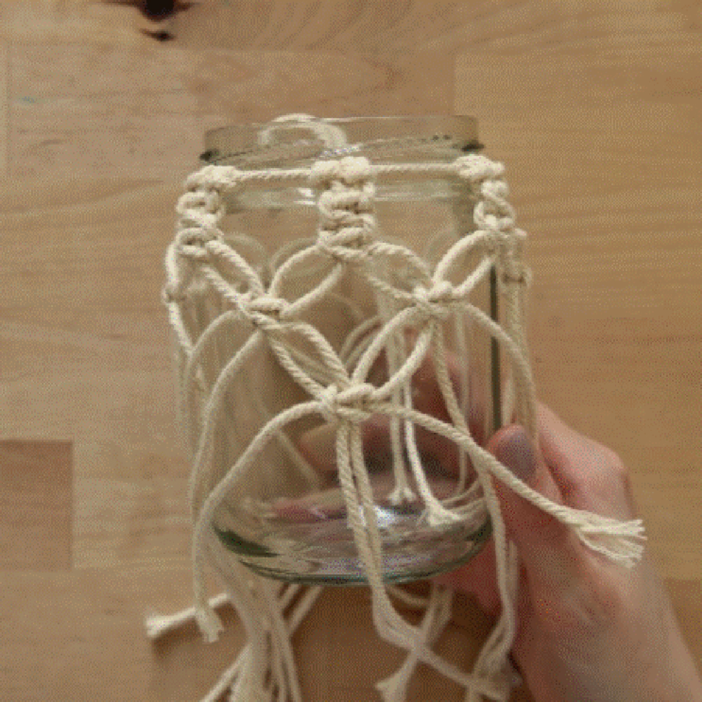 Une main tient un bocal décoré de macramé sur une table en bois.
