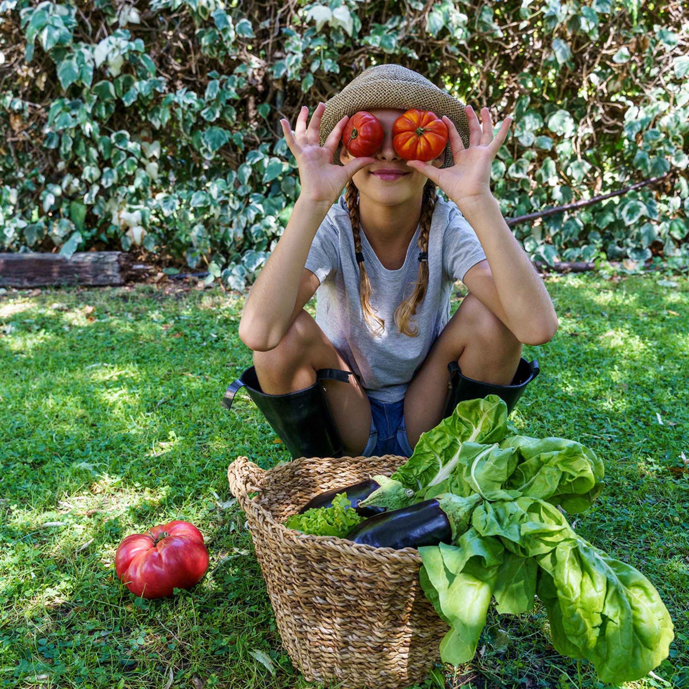 Une personne tient des tomates devant son visage, assise dans le jardin à côté d'un panier de légumes.