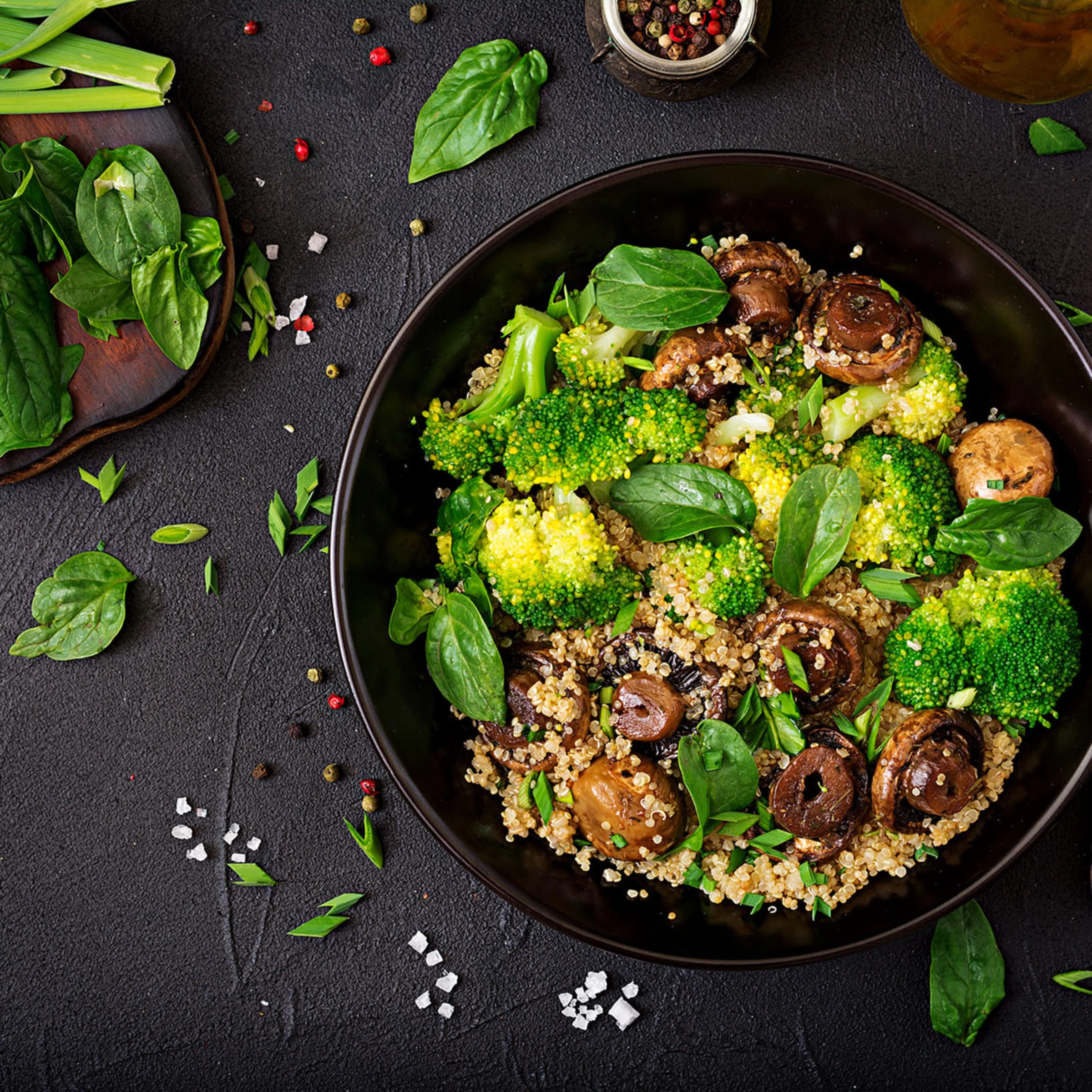 Assiette noire avec salade de quinoa, brocoli, champignons et feuilles de basilic sur table sombre.