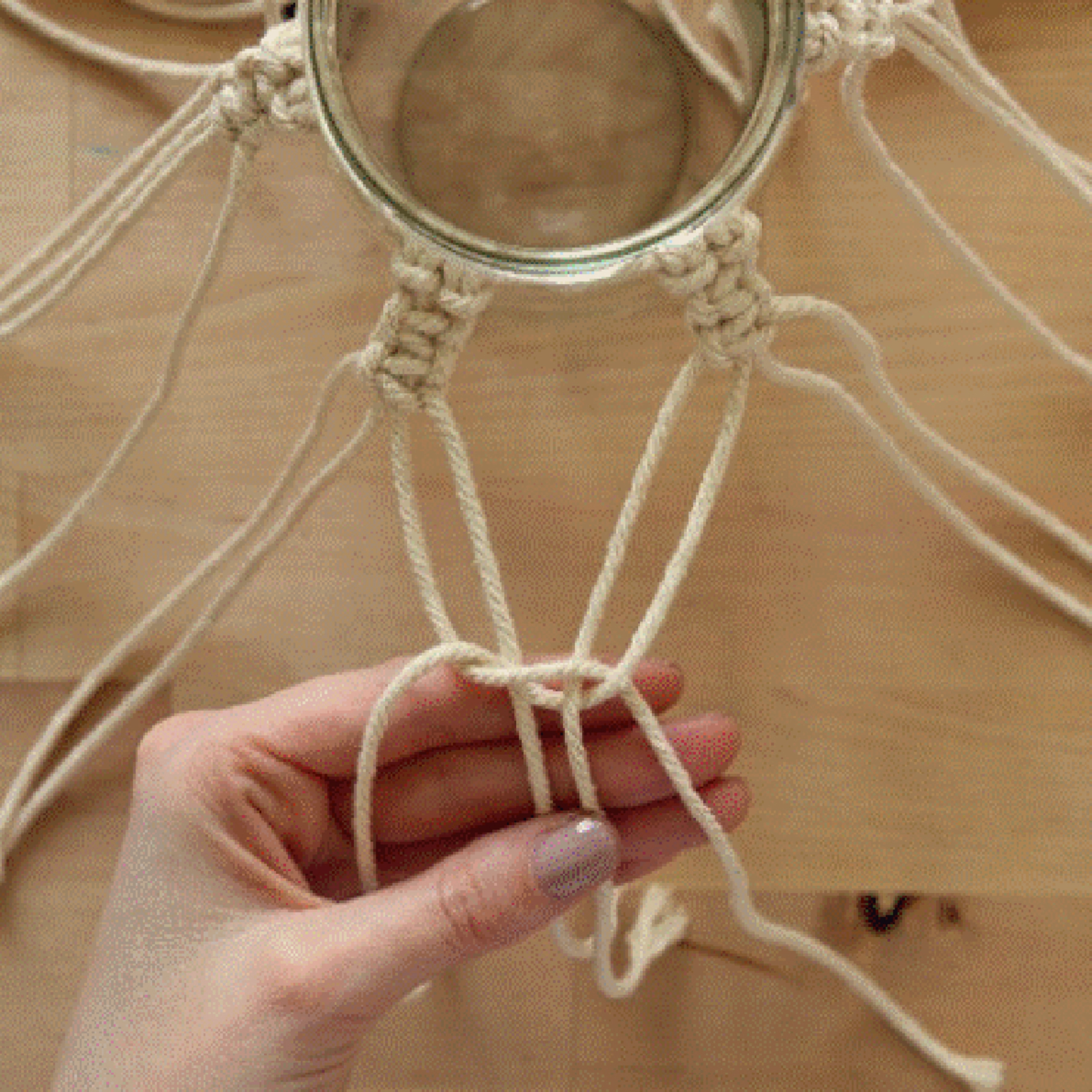 Une main noue des cordes de macramé autour d'un bocal sur une table en bois.