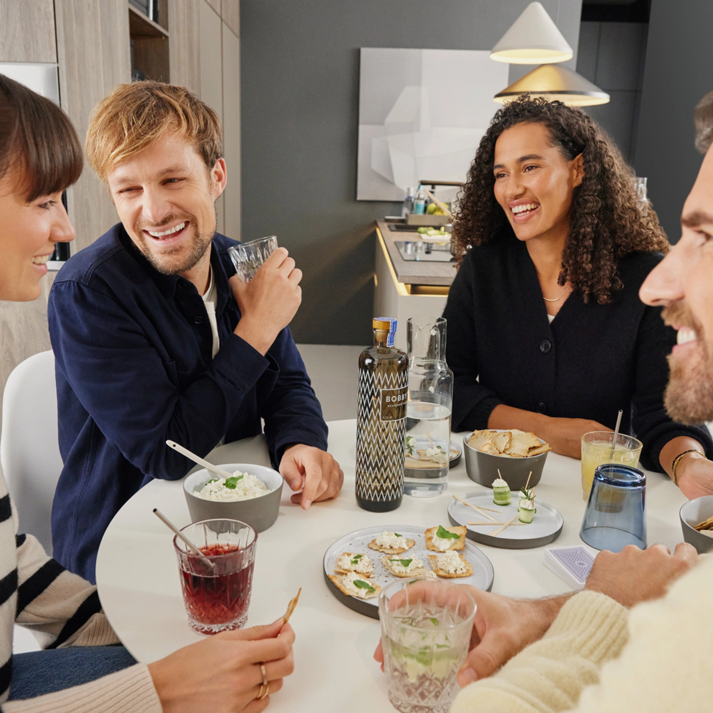 Quatre personnes assises joyeusement autour d'une table avec boissons et snacks dans un salon moderne. Nolte Living.
