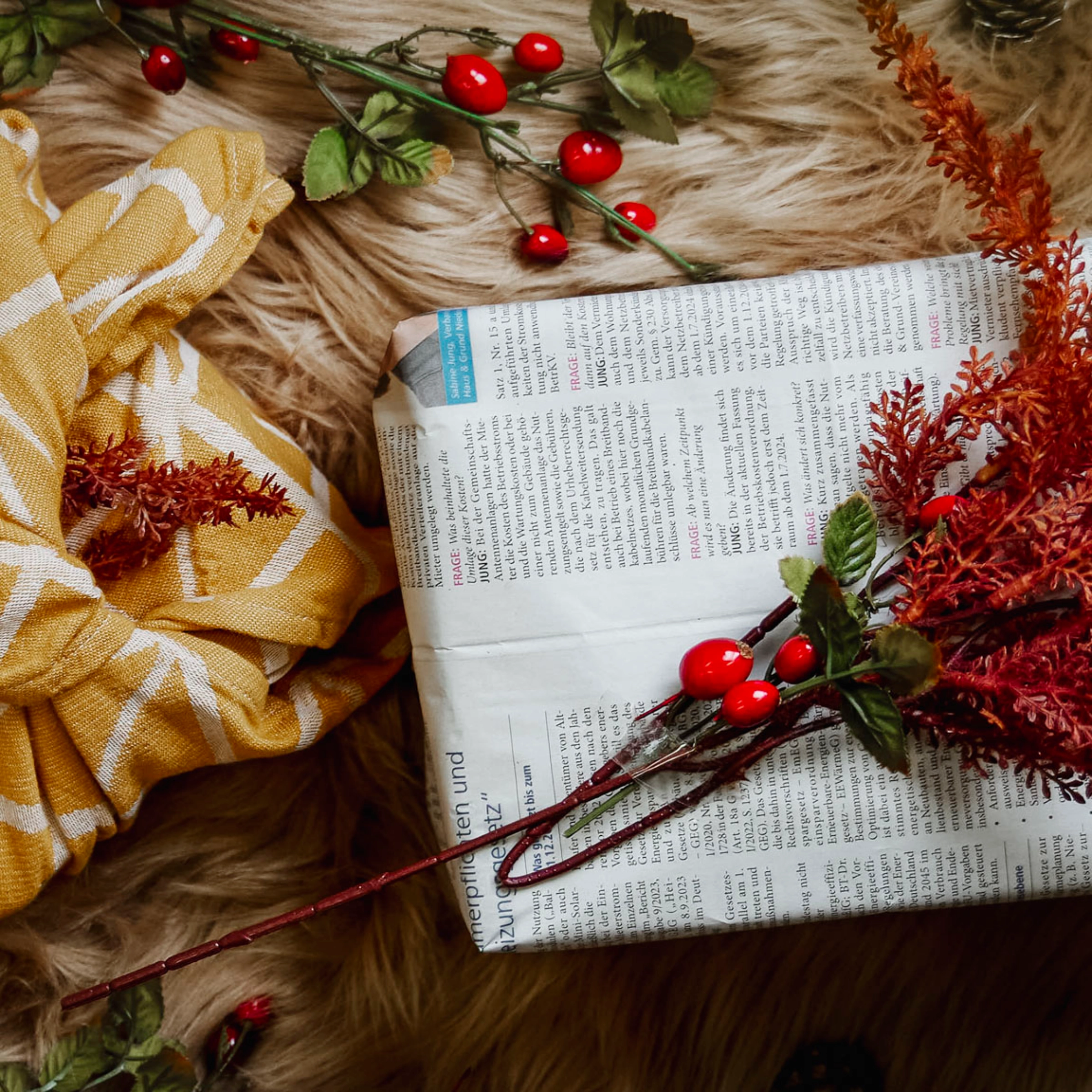 Ein mit Zeitungspapier und roten Beeren dekoriertes Geschenk liegt auf einem Fell neben einem gelben Tuch.