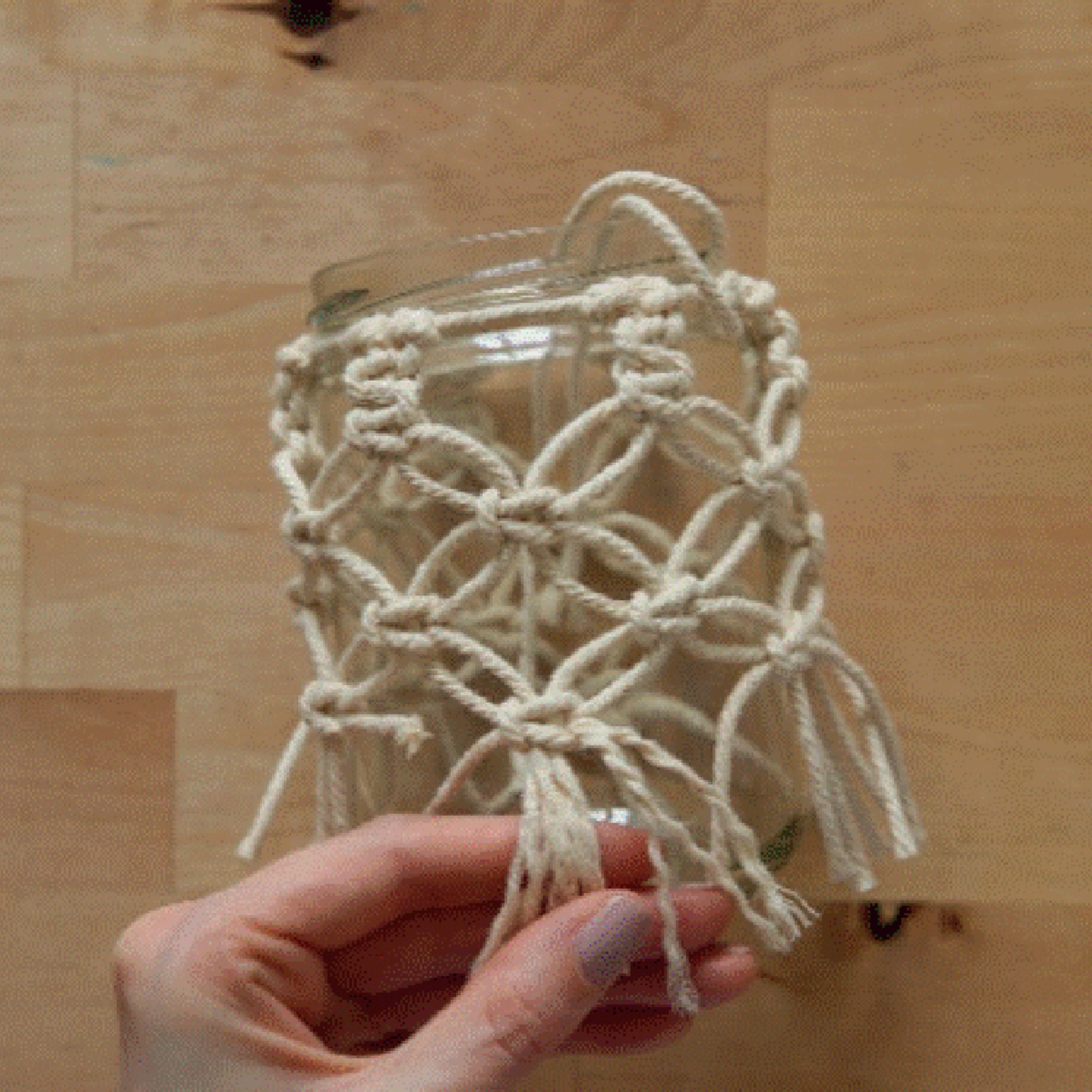 Une main tient un verre décoré de macramé sur une table en bois.