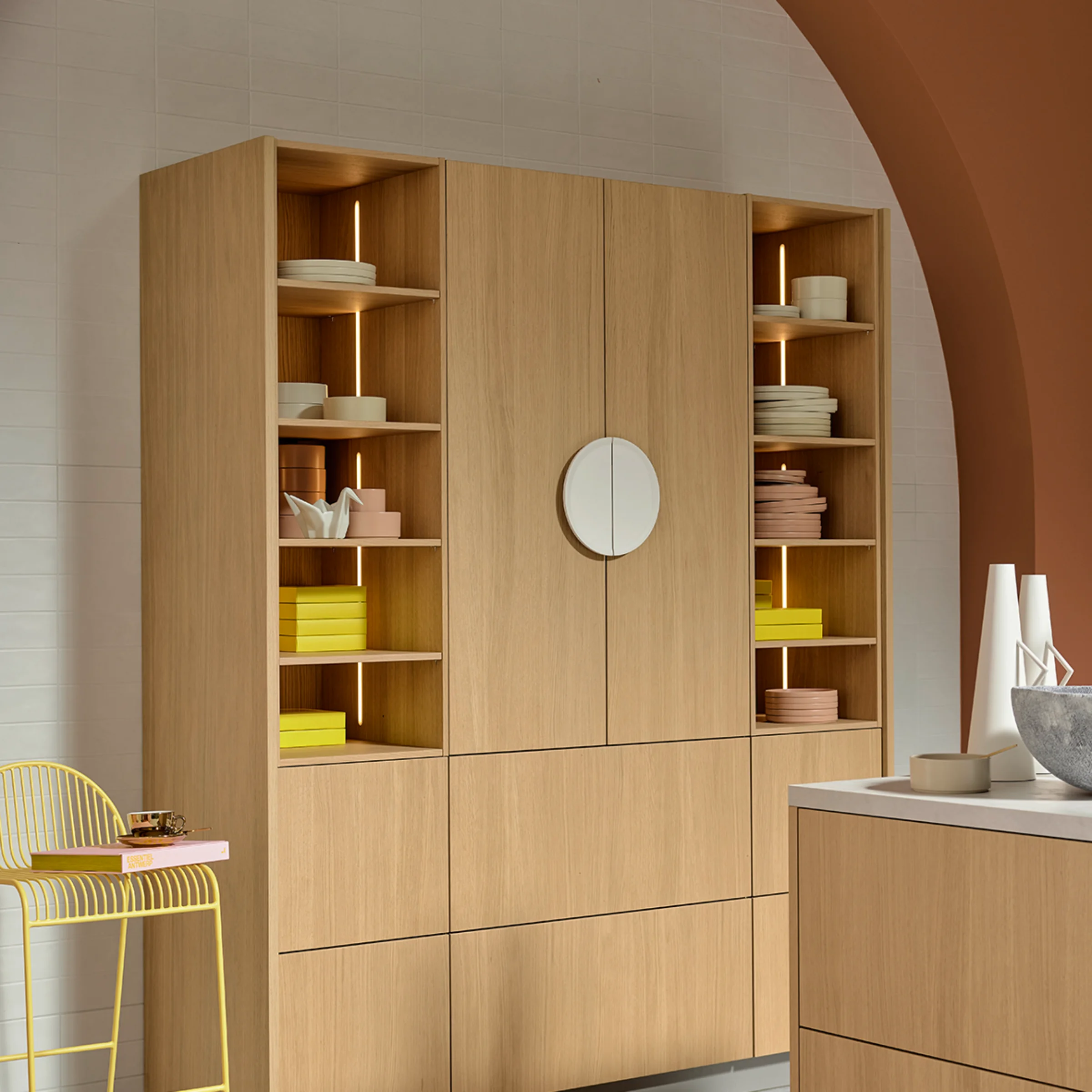 Moderner Hochschrank in Eiche Bianco mit offenen Regalfächern und integrierter Beleuchtung unter rundem Bogen in Terrakottaton. Warmes Farbkonzept mit gelben Akzenten und minimalistischem Design von nolteneo.